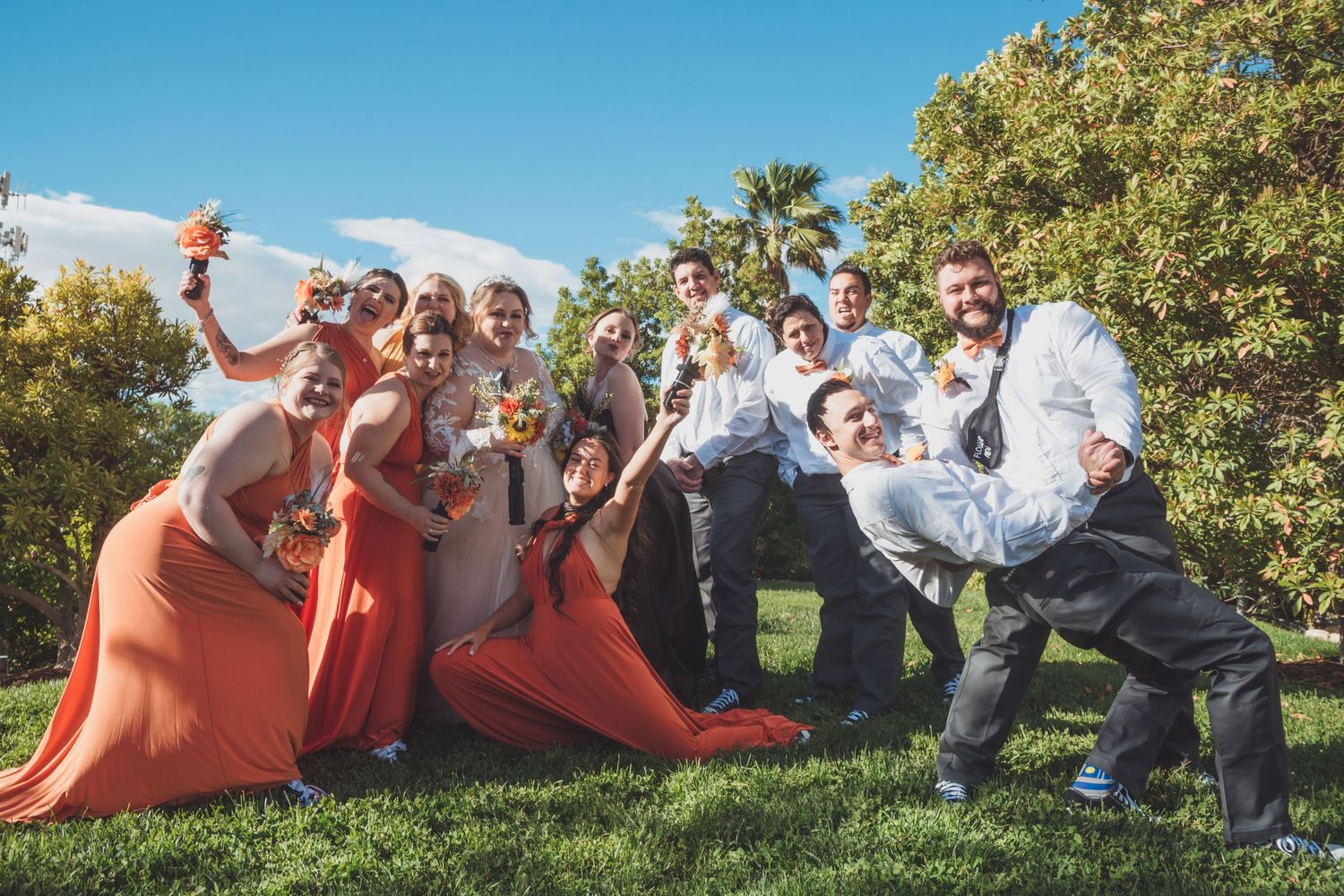 Wedding party posing playfully on green grass, bridesmaids in coral dresses with groomsmen, trees and blue sky behind.