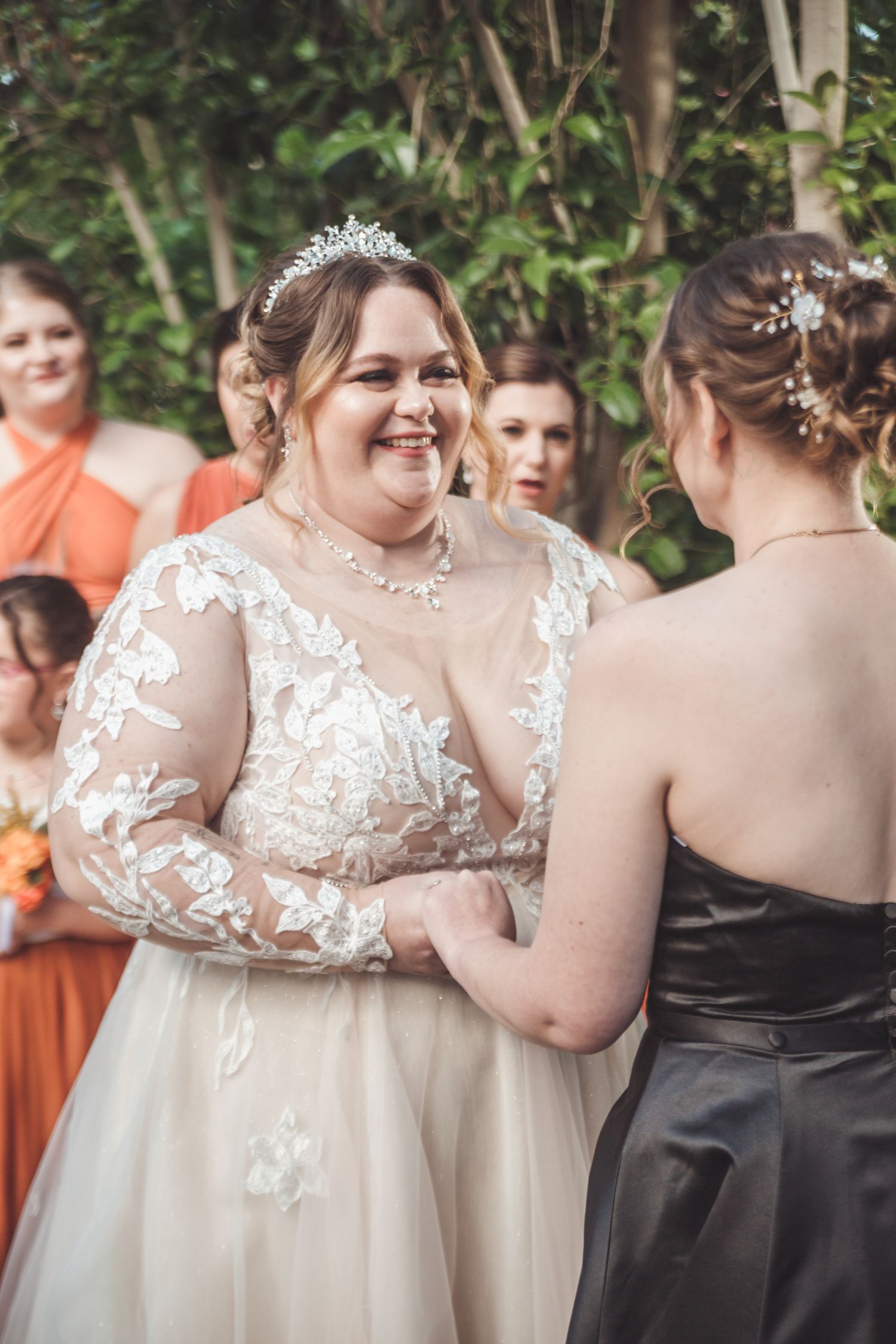 Two brides hold hands during outdoor wedding ceremony, one in white lace gown with tiara, one in black dress.