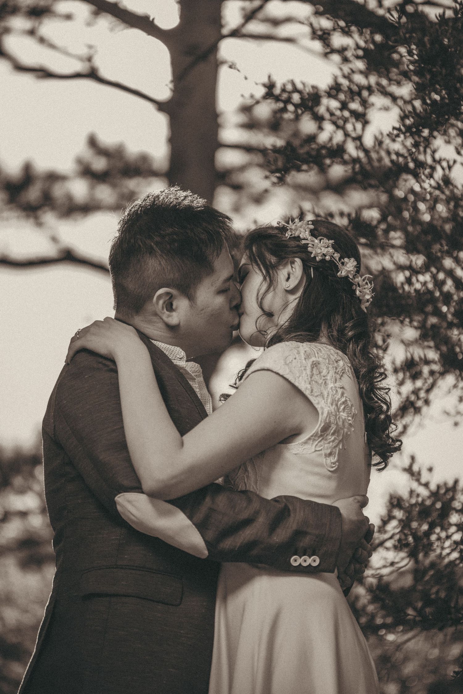 Bride and groom sharing a romantic kiss outdoors, with the bride wearing a floral crown, in a sepia-toned wedding photo.