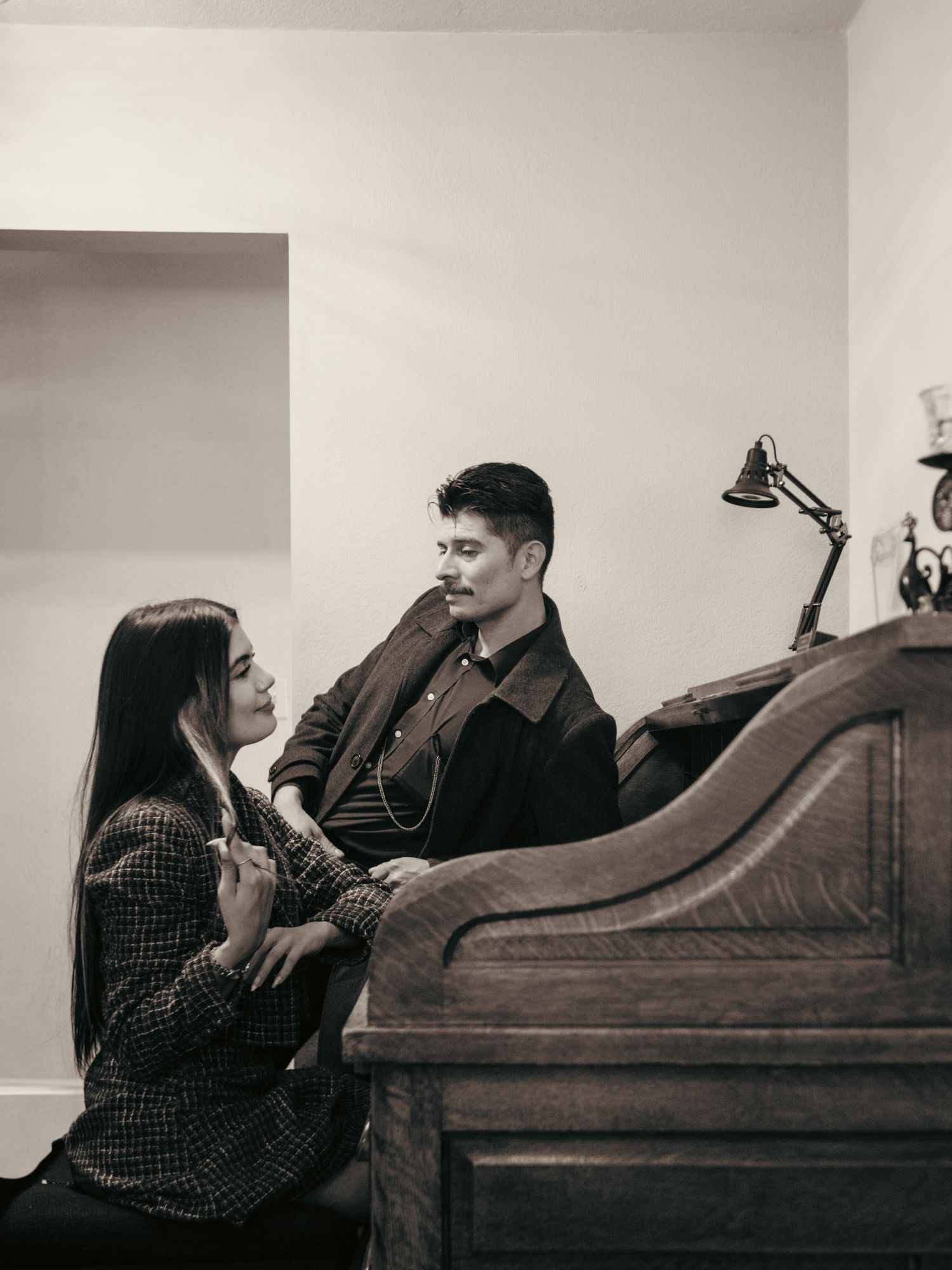 A couple shares an intimate moment beside a vintage wooden piano in a softly lit room, captured in black and white.
