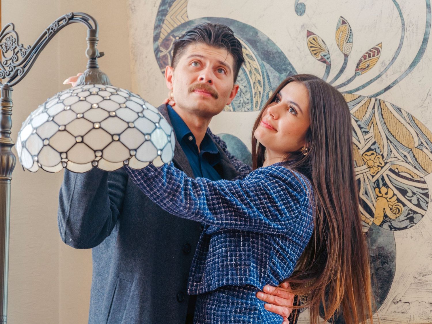 A couple poses near a Tiffany-style lamp against a decorative wall, both wearing blue plaid shirts.