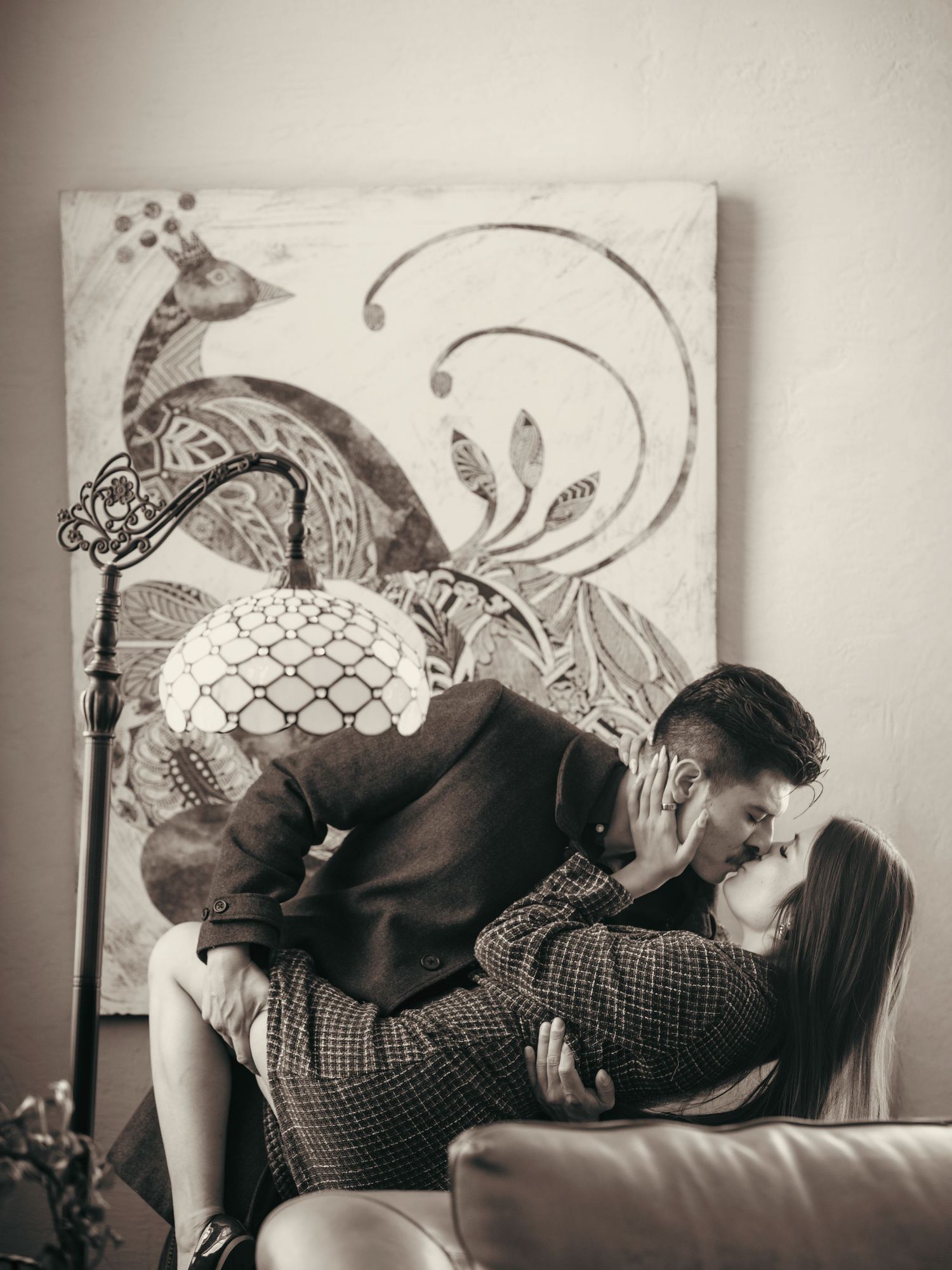 Black and white photo of a romantic couple kissing in a cozy room with a decorative peacock painting backdrop.