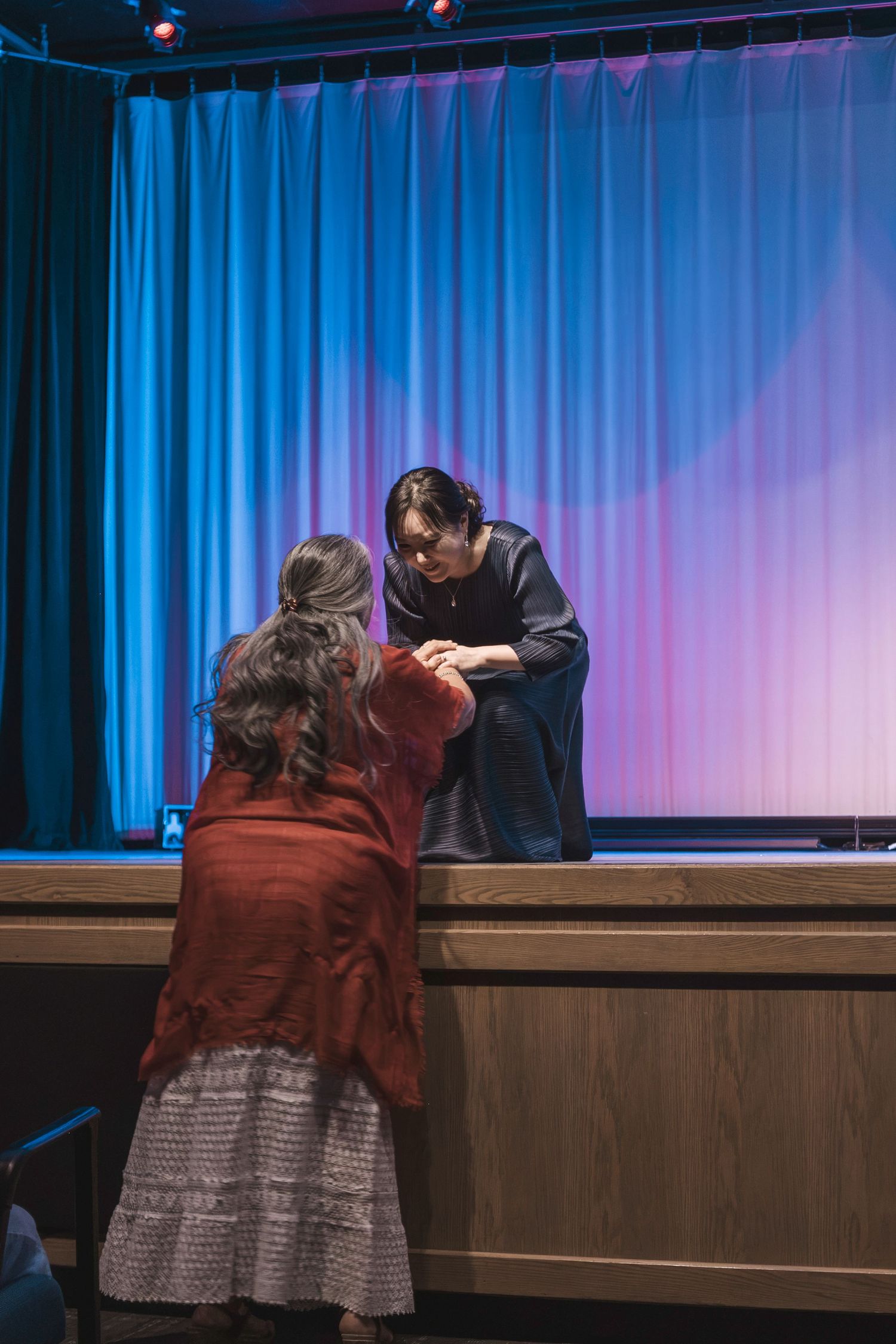 Two people converse near a stage with blue and pink curtains in a theater setting.