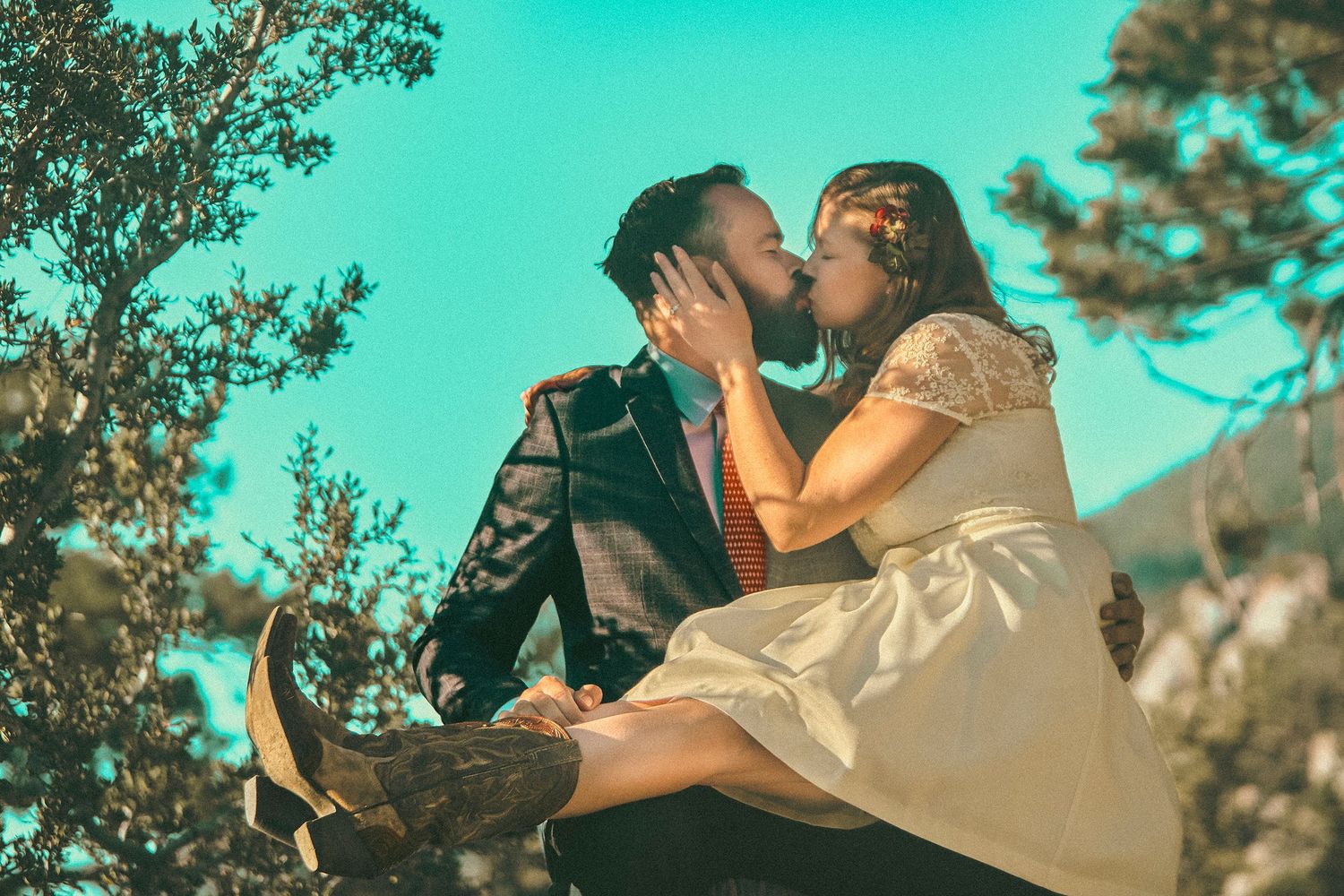 A couple shares a romantic kiss outdoors, with the groom holding the bride in his arms surrounded by trees.