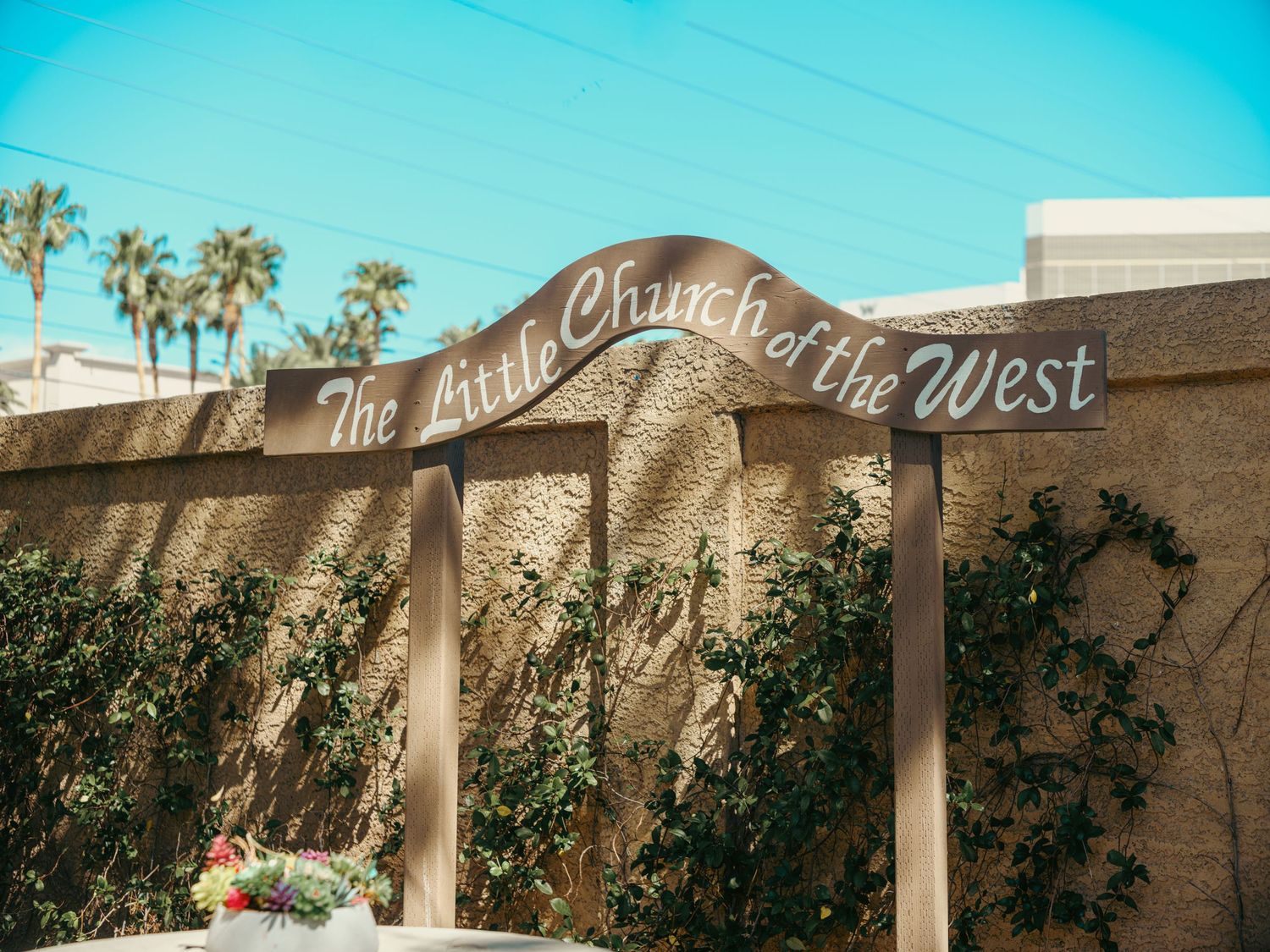 Rustic wooden sign reading The Little Church of the West mounted on a stucco wall with ivy under a clear blue sky.