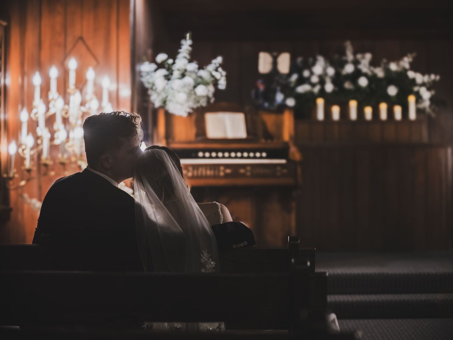 Bride and groom sitting together in a dimly lit church pew, surrounded by candlelight and white floral arrangements.