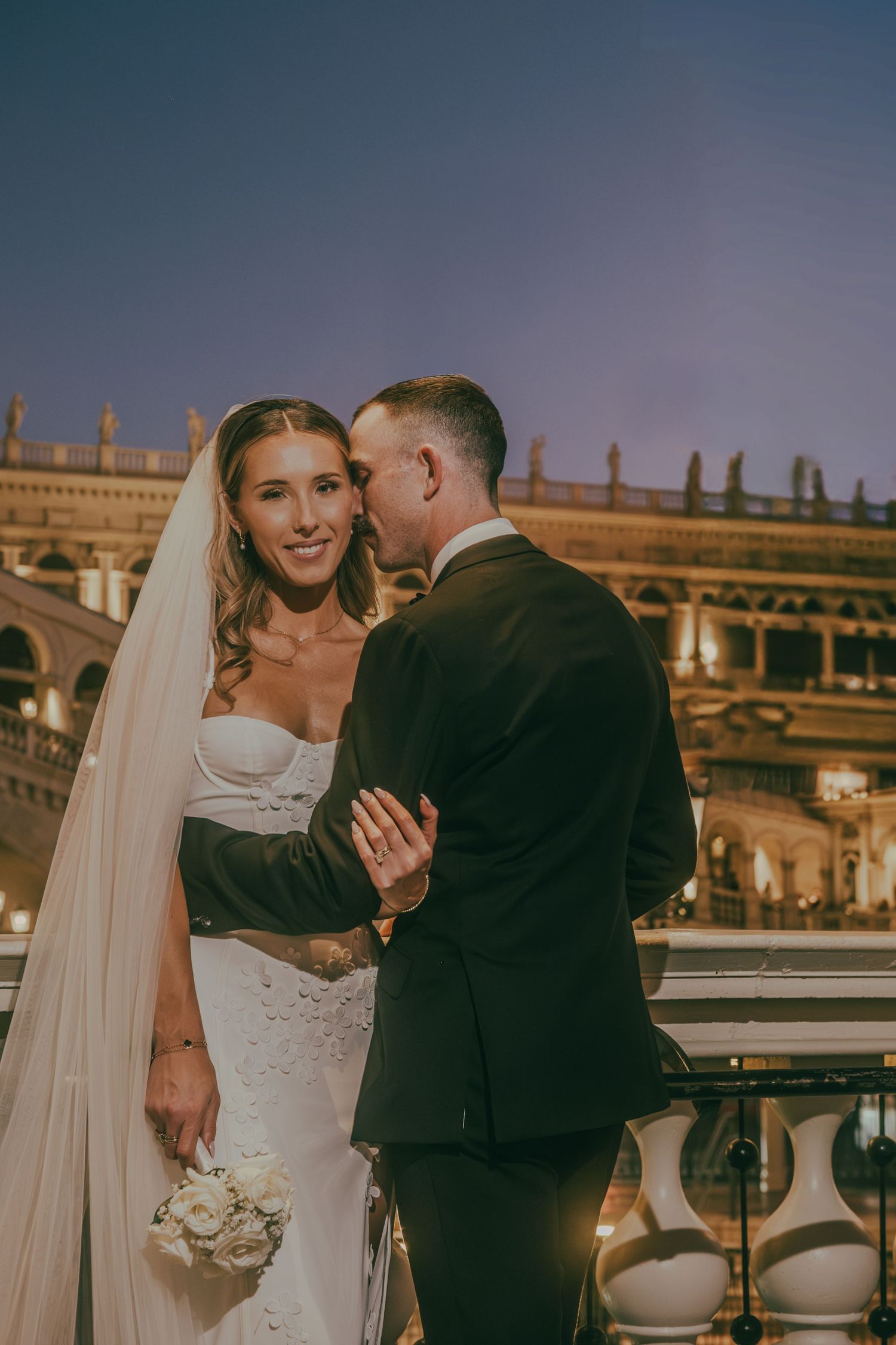 Smiling bride in a white gown holds flowers as groom whispers to her against a glowing Las Vegas backdrop.