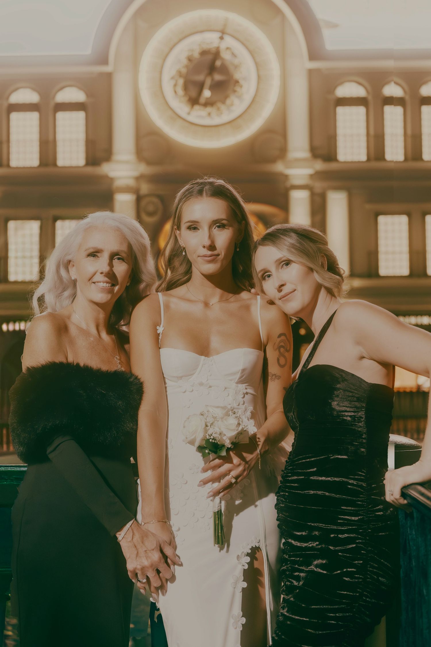 Bride in white gown poses with two women in black dresses before ornate clock at Las Vegas casino.