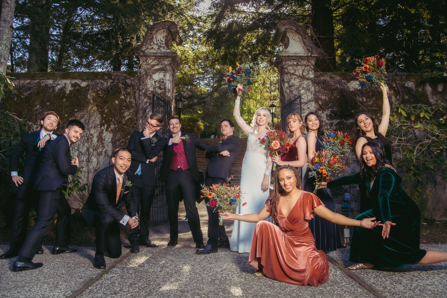 Wedding party celebrates playfully at stone gate entrance, featuring bridesmaids in rich jewel-toned velvet gowns.