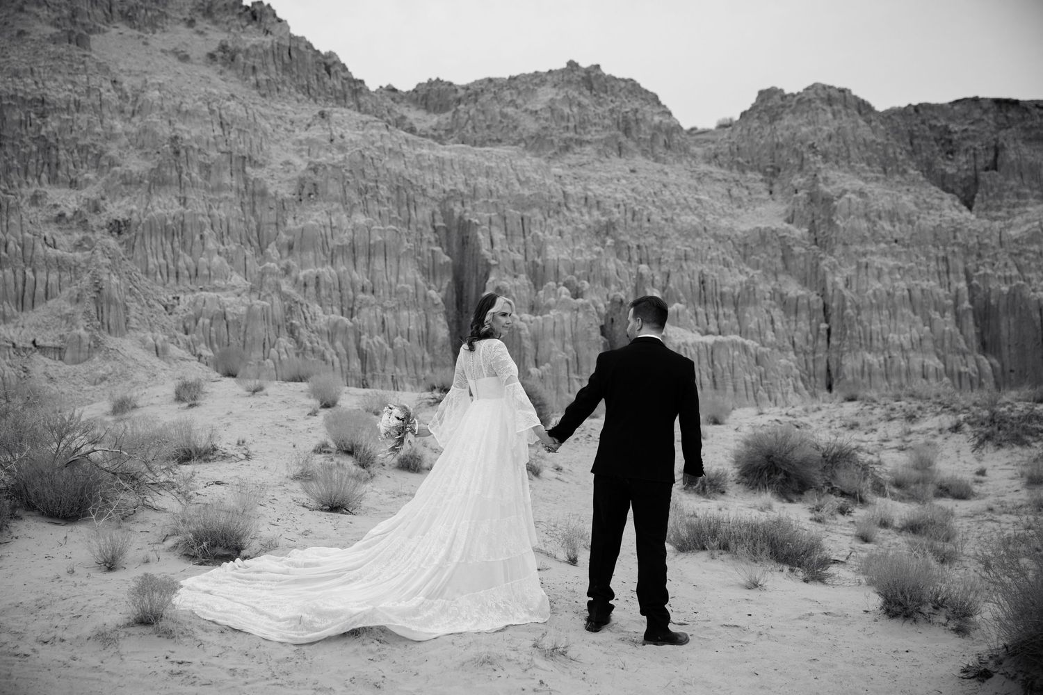 Black and white photo of bride and groom holding hands facing dramatic desert rock formations.