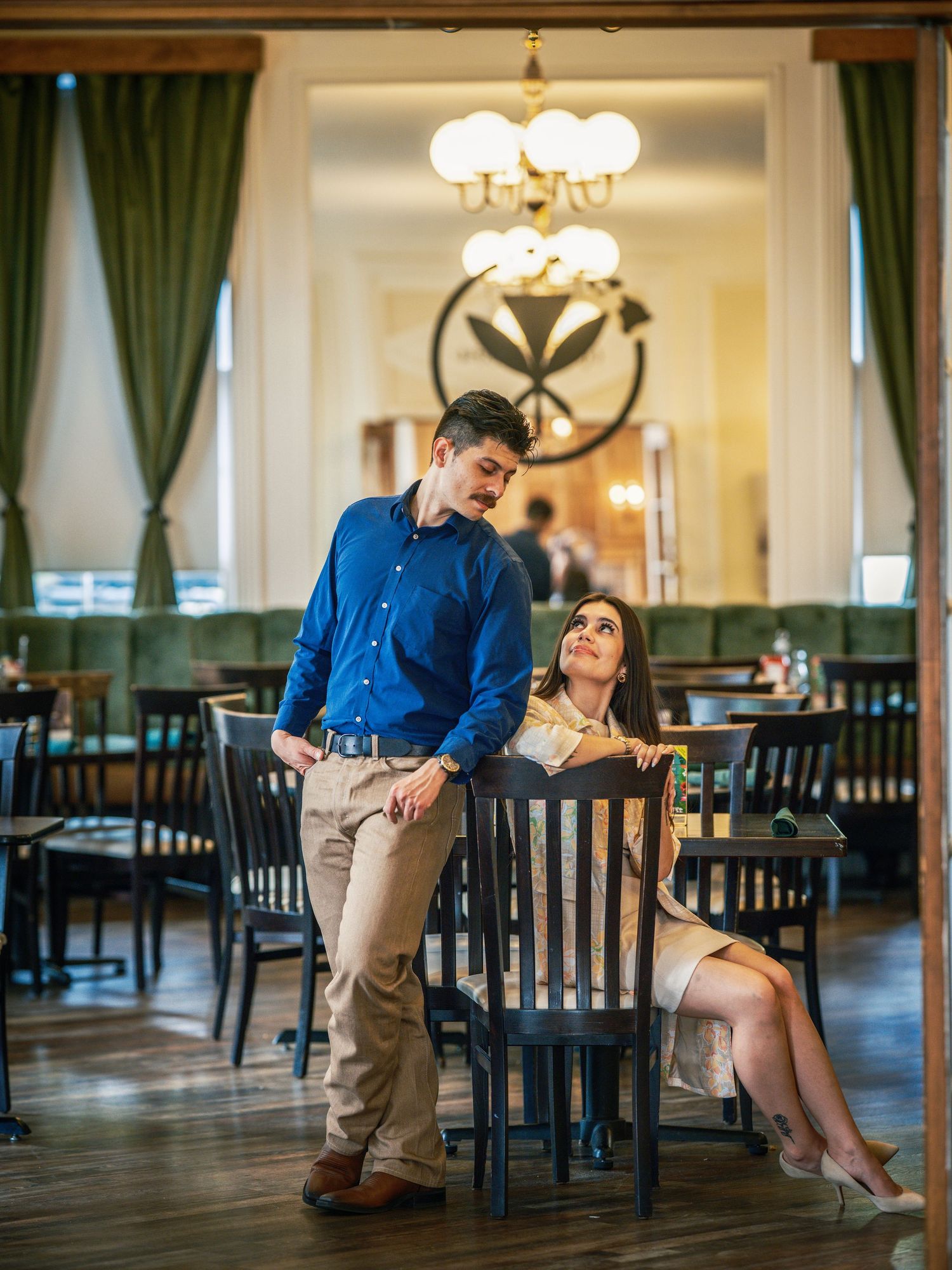 Couple posing in elegant restaurant interior with chandeliers, man in blue shirt standing by woman seated at dark wood chair.