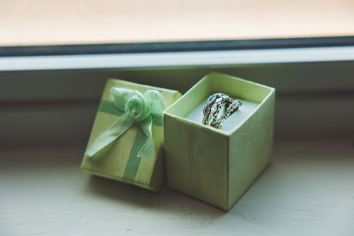 A silver ring displayed in an open green gift box with a ribbon bow, placed on a white windowsill.