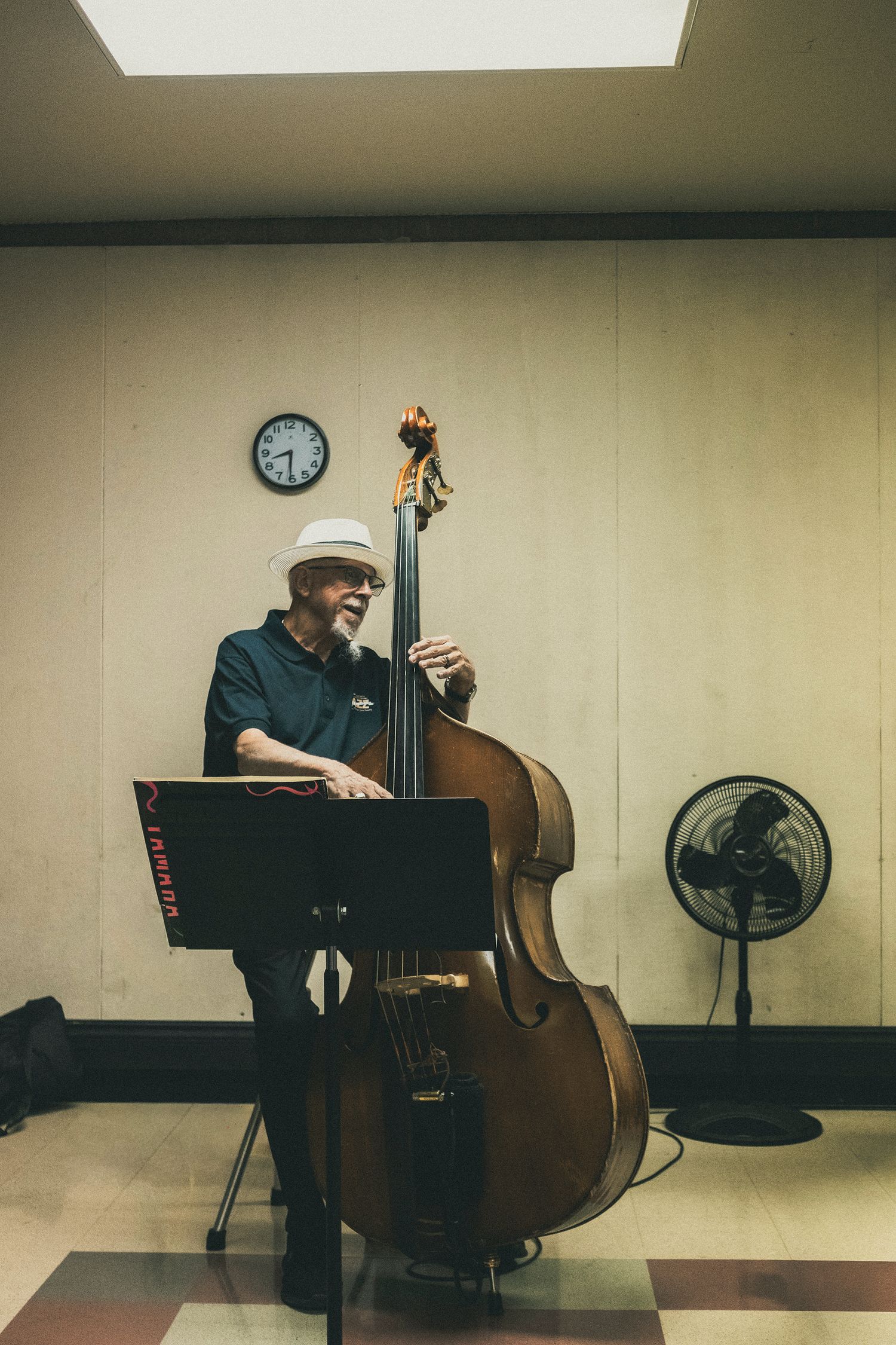 Elderly musician playing a large double bass in a room with a wall clock and fan.