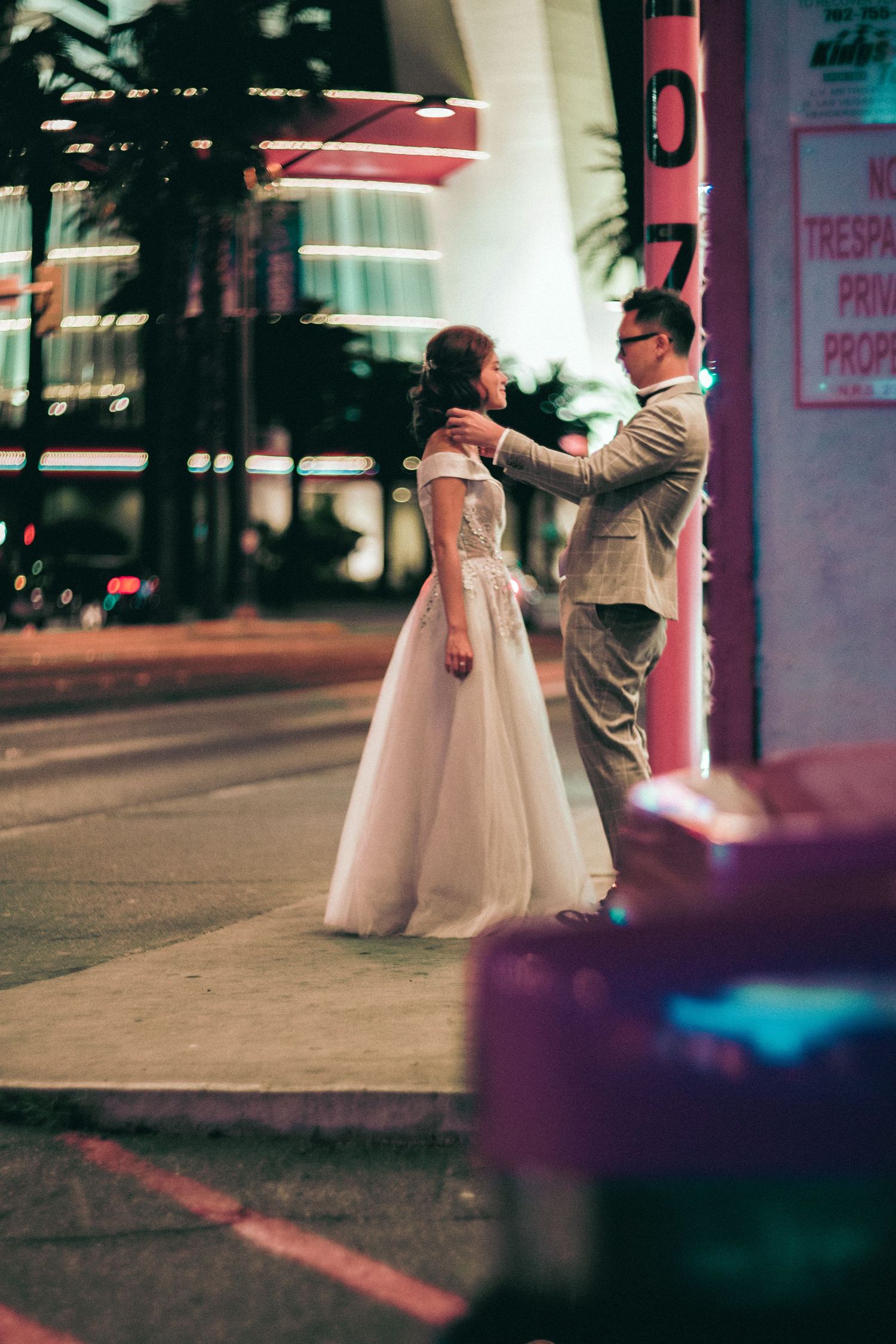 Couple in wedding attire standing on a city sidewalk at night, groom adjusting bride's dress near neon-lit storefront.