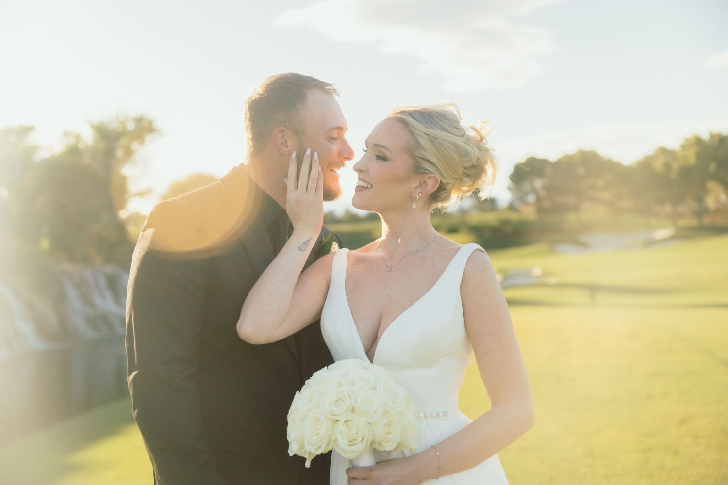 Bride holding white bouquet tenderly touches grooms face in golden hour light.