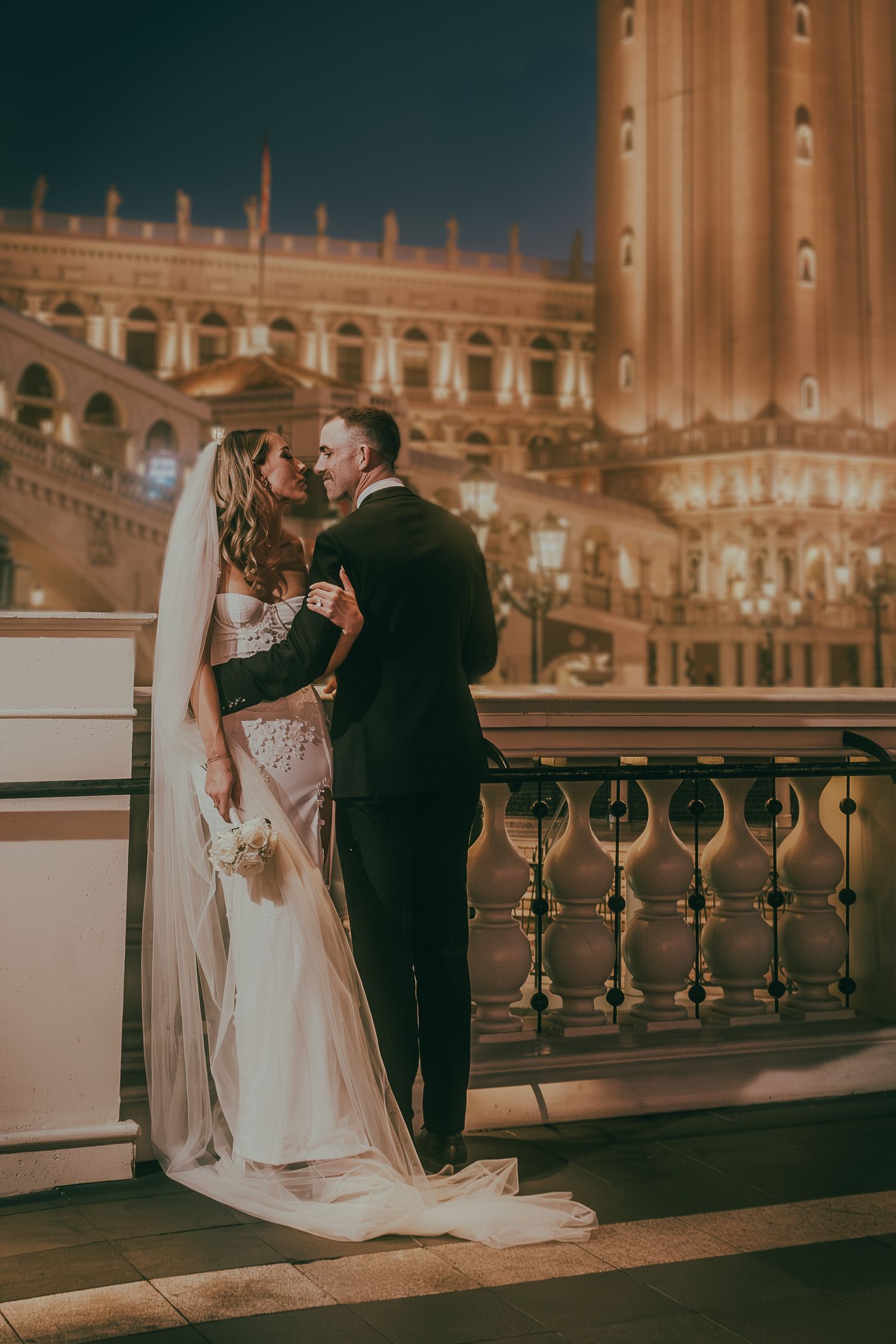 Bride and groom share a romantic kiss on a balcony with a glowing Las Vegas cityscape at night.