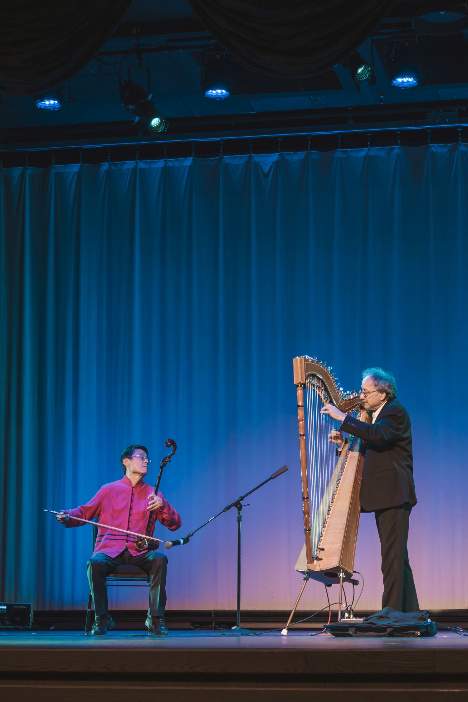 Two musicians perform on stage, one playing an erhu and one playing a harp, against a blue curtain backdrop.