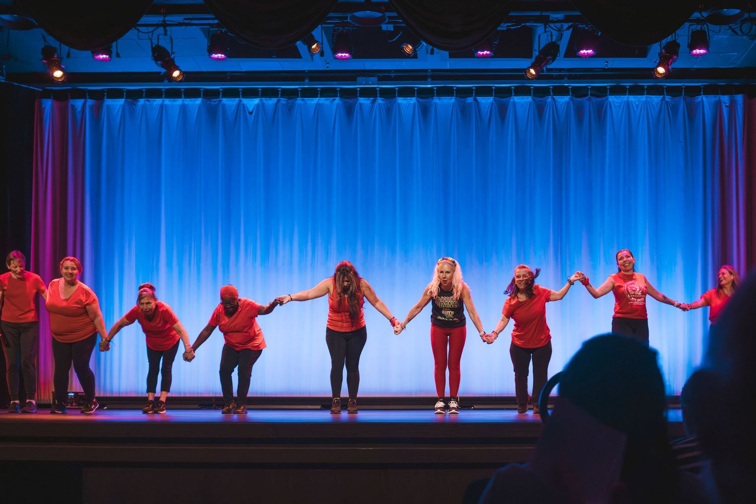 Performers in red shirts hold hands on a blue-lit stage during a theatrical curtain call.