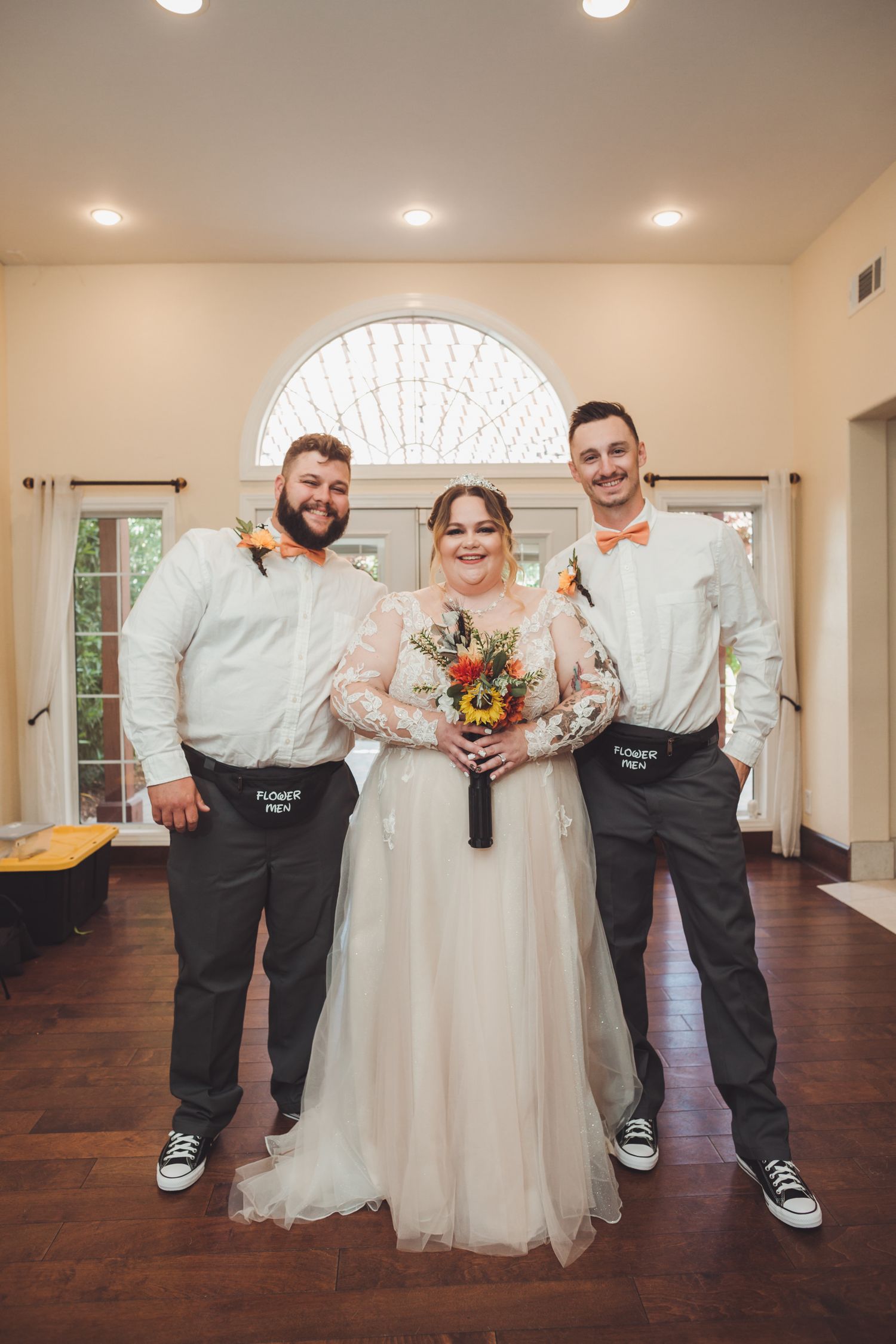 Bride in lace gown poses with two groomsmen in orange bowties and black aprons indoors.