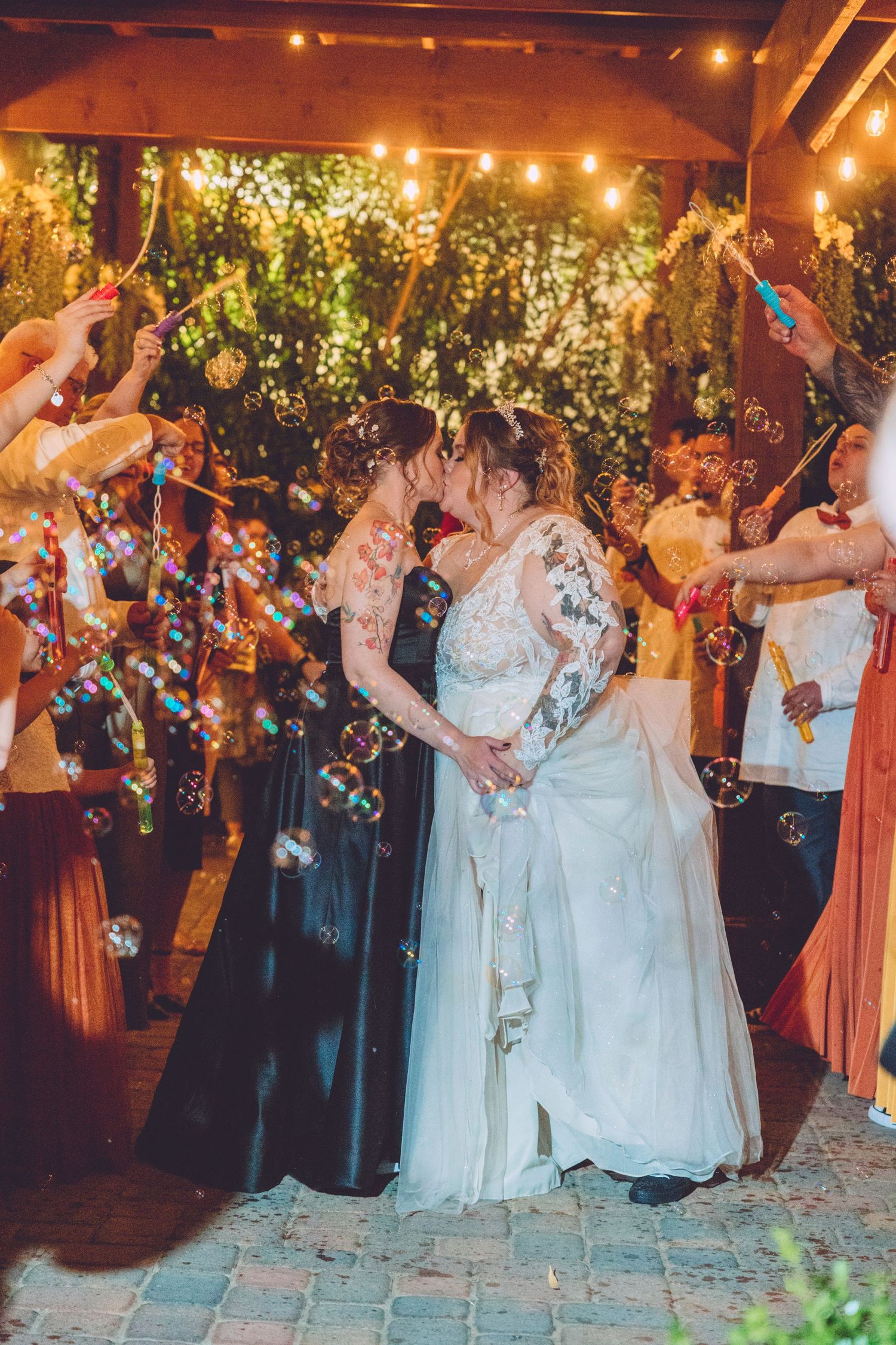 Two brides share a kiss surrounded by guests blowing colorful bubbles at an outdoor evening wedding celebration.