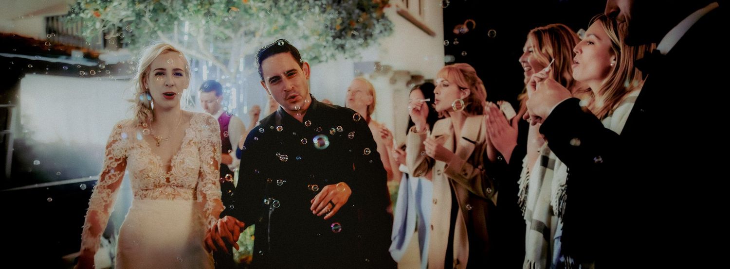 A bride and groom walk through a confetti shower surrounded by cheering wedding guests at an outdoor evening celebration.