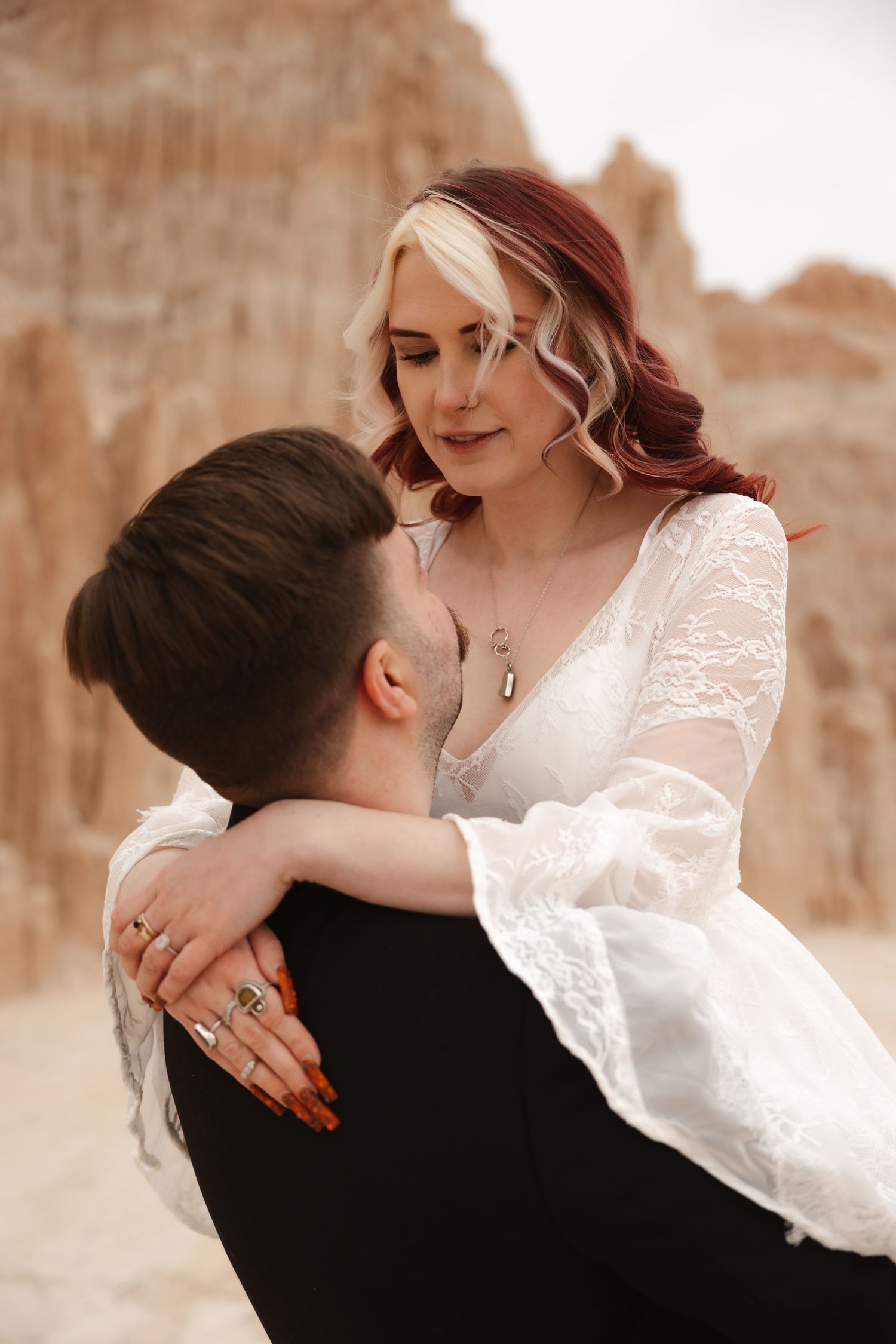 Bride with red and blonde hair gazes down at groom while he lifts her, wearing white lace boho wedding dress.