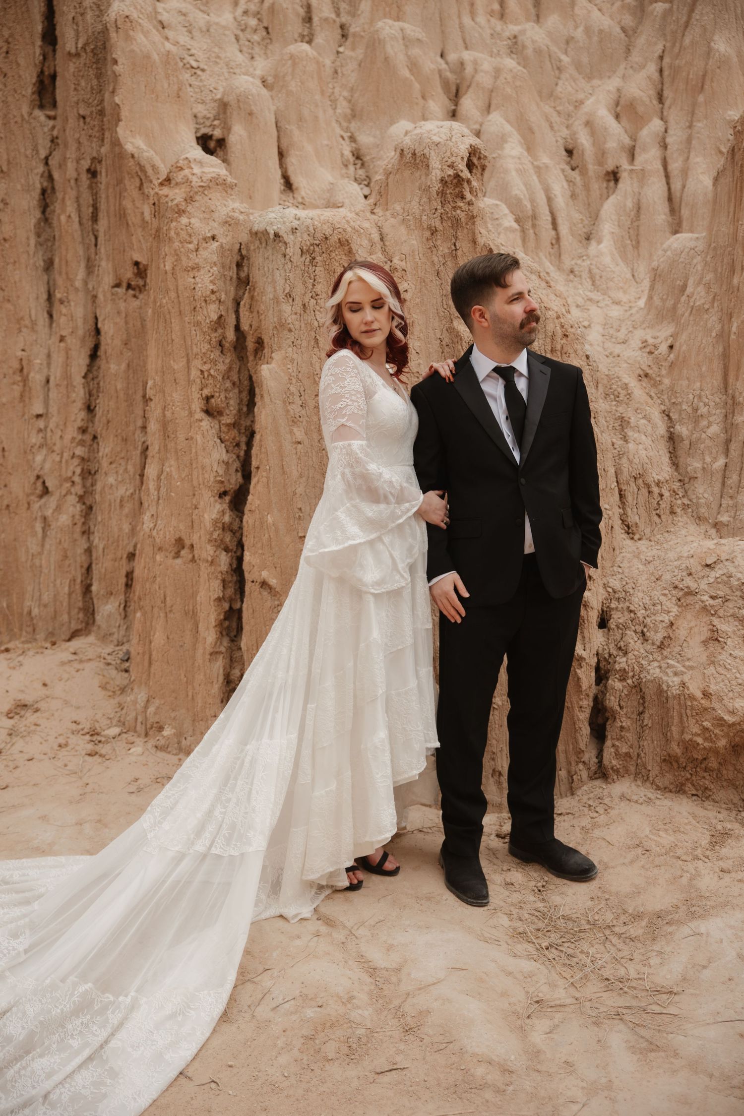 Bride in flowing white lace gown and groom stand together against textured sandy rock backdrop.