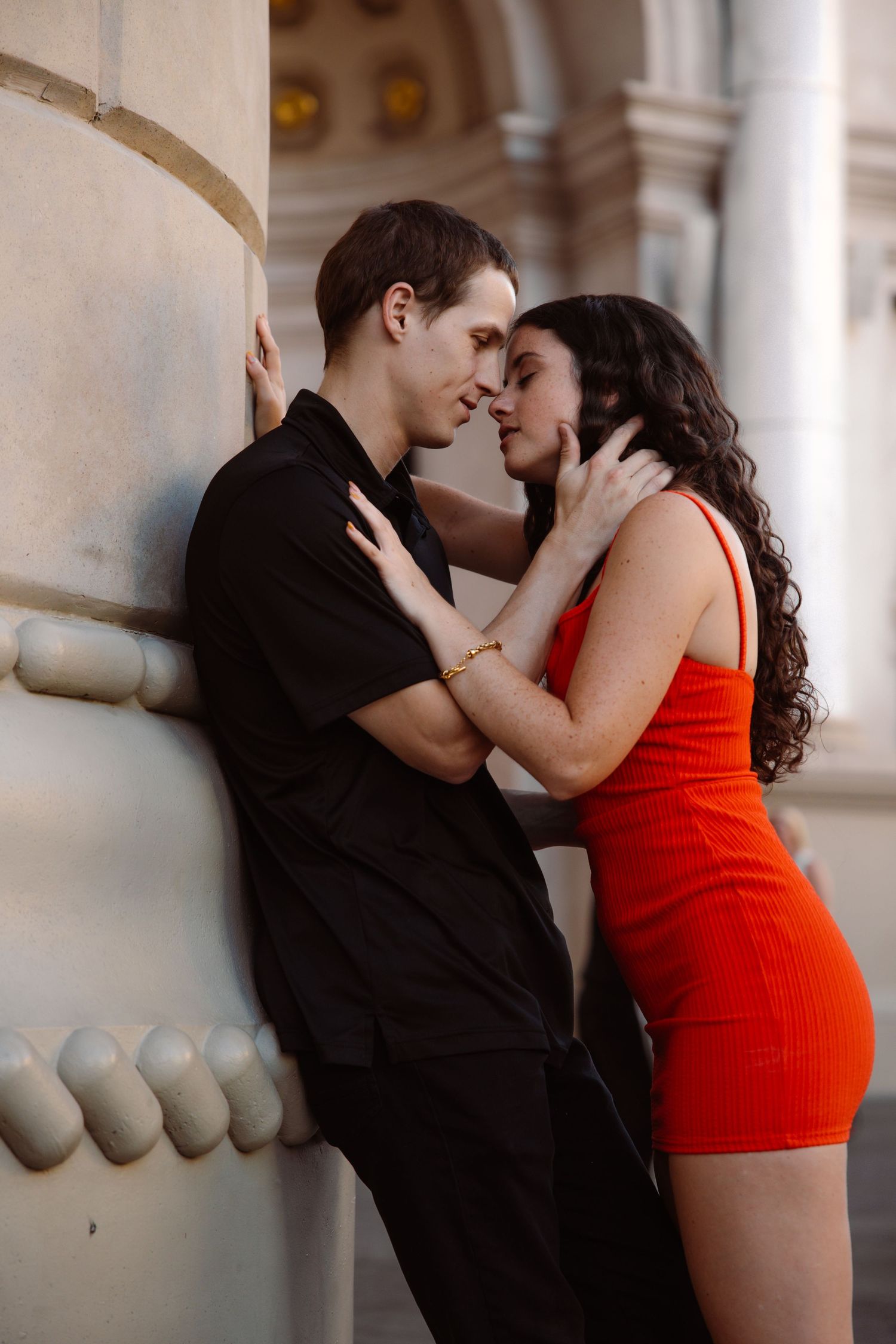 Romantic couple nearly kissing against classical stone column, woman in red dress, man in black shirt.