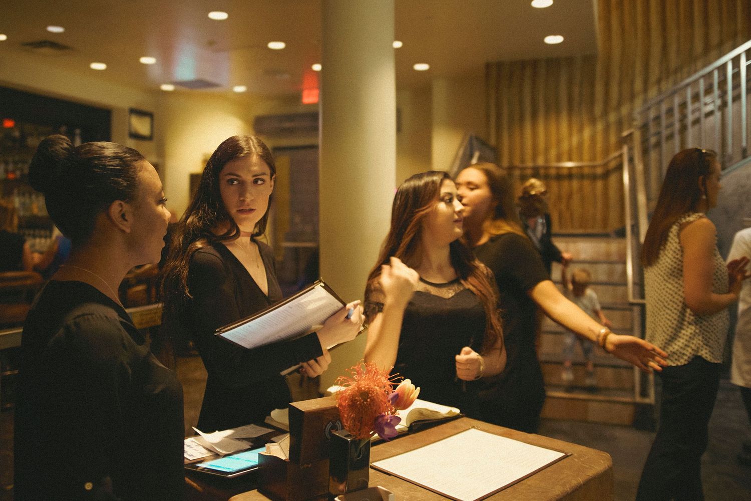 Women in black attire gather at a dimly lit venue reception desk, reviewing papers at an evening event.