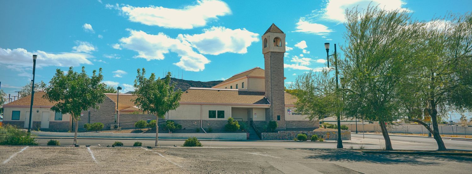 A church with a clock tower surrounded by trees under a blue sky with clouds in a desert town.
