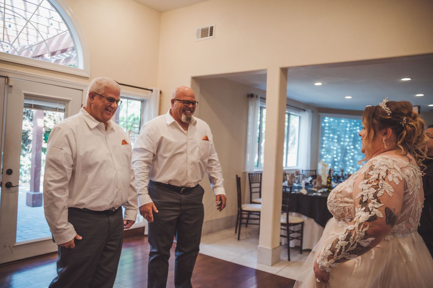 Bride in lace gown smiles at two men in white shirts during an elegant indoor wedding venue celebration.