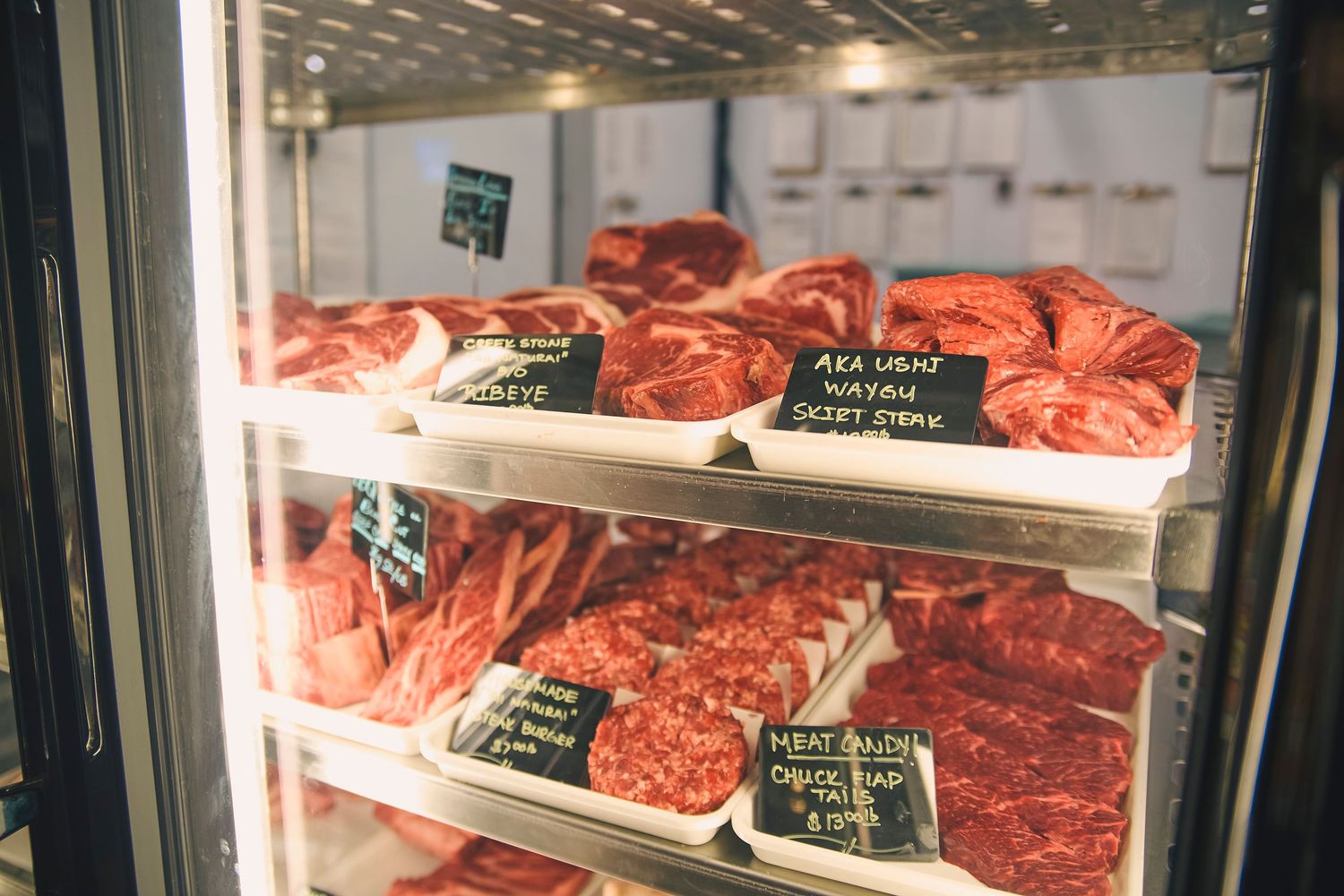 Fresh cuts of premium beef displayed on refrigerated shelves with chalkboard price tags in a butcher shop.