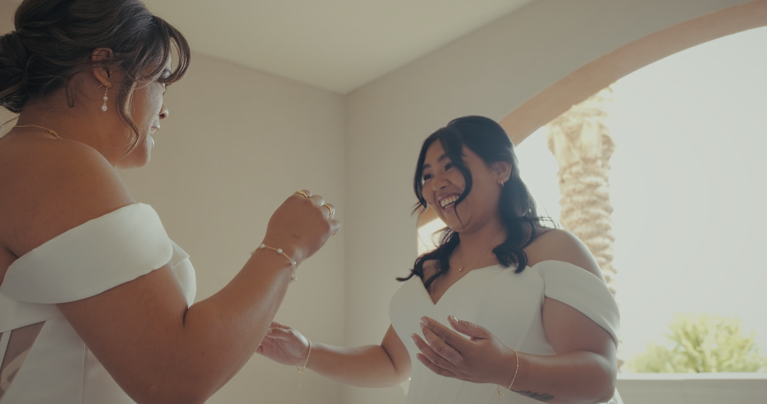 Two women in white off-shoulder dresses laugh and hold hands in sunlit space.