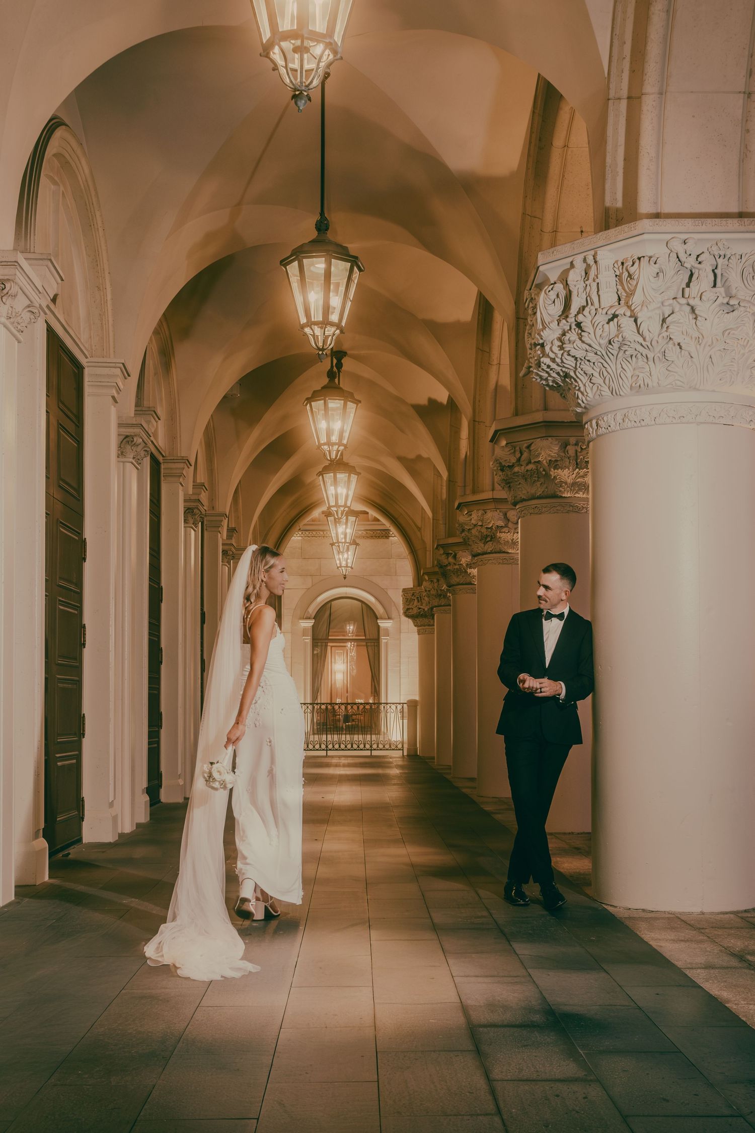Bride in white veil gown and groom in tuxedo share glance in elegant lantern-lit corridor.