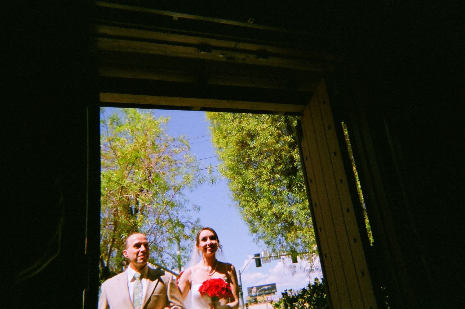 Bride and groom photographed through a doorway framed by lush green trees and blue sky on their wedding day.