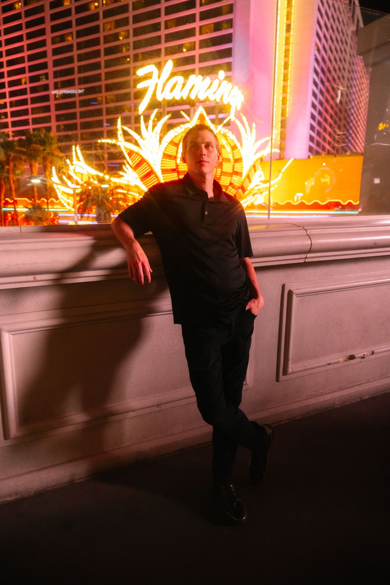 Man in black outfit leans against wall in front of glowing Flamingo hotel neon sign in Las Vegas.