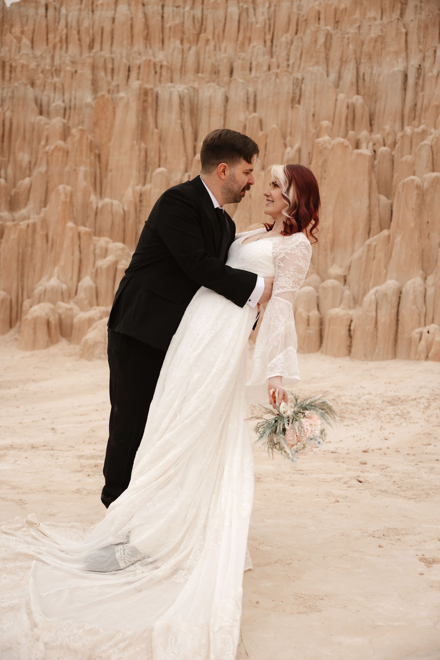 Groom dips bride in white lace wedding gown before tall sandy rock spires, she holds a boho floral bouquet.