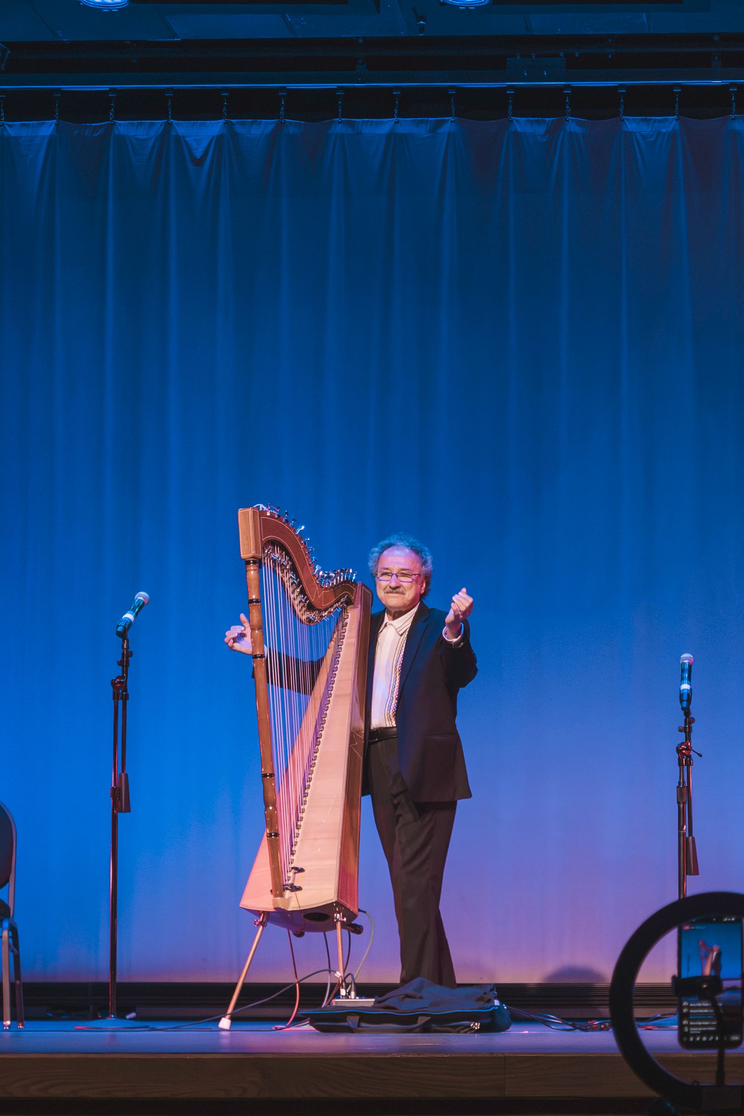 Man in tuxedo stands on stage beside a wooden harp under blue stage lighting at a live performance.