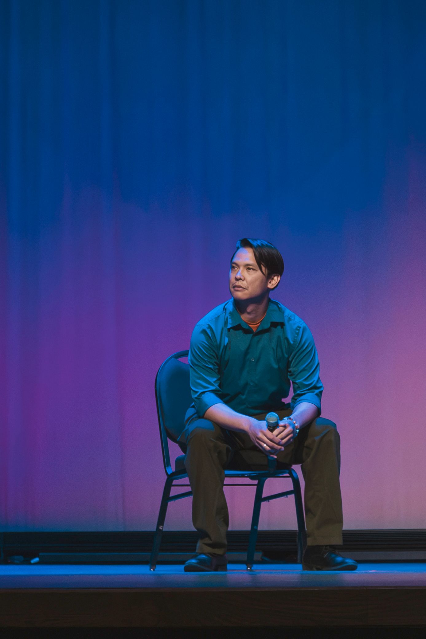 Man sitting on stage chair holding microphone under blue and pink lighting during a performance.