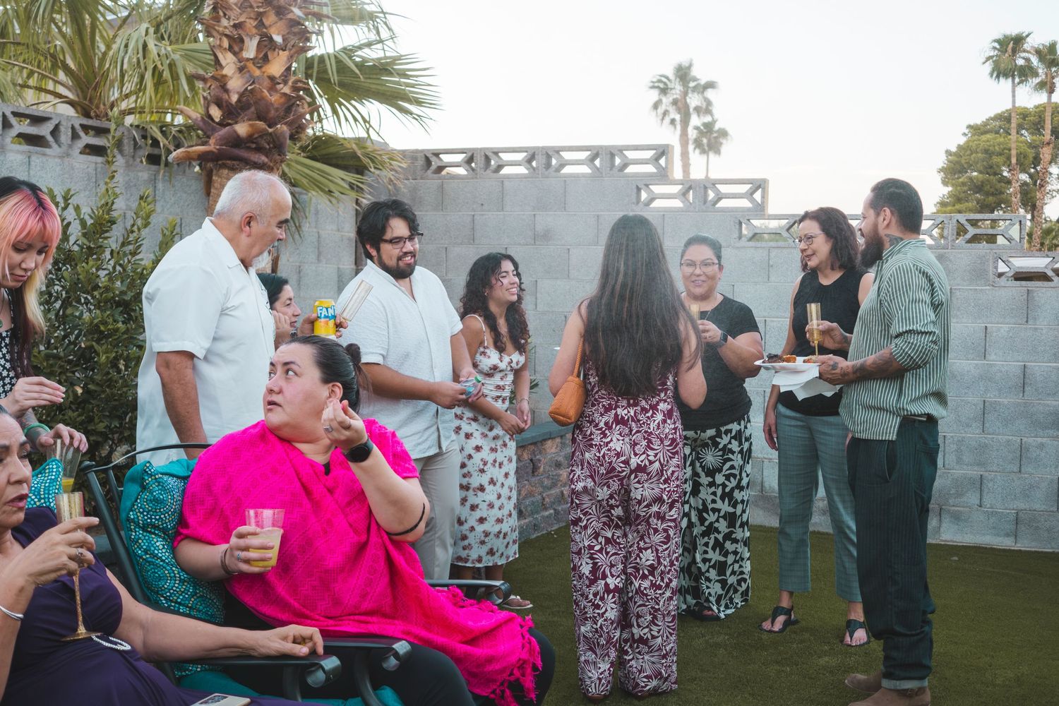 Diverse group of people socializing outdoors at a casual garden party near a stone wall with palm trees.