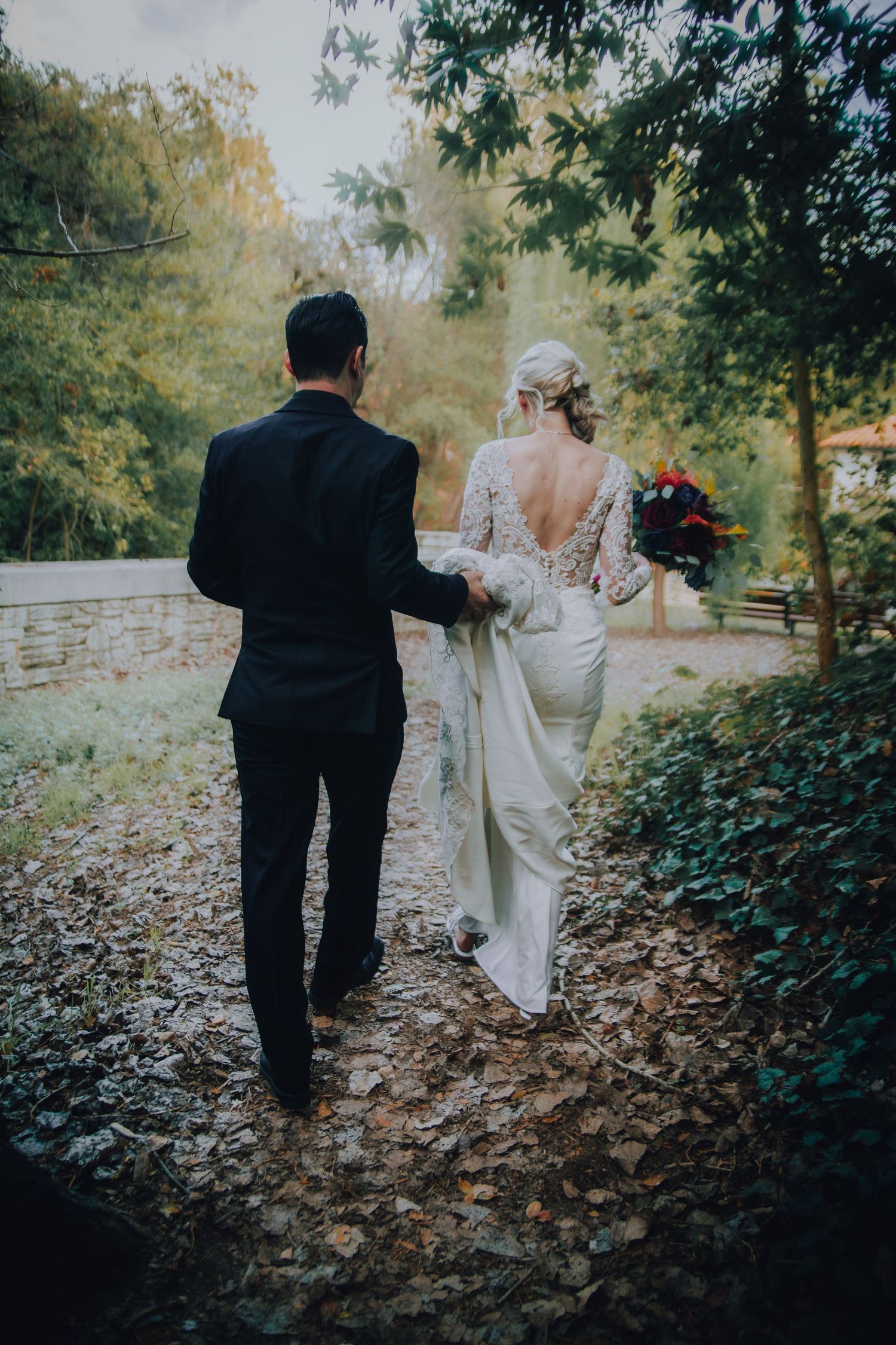 Bride and groom walking hand in hand through a leafy outdoor path, bride wearing a lace backless wedding gown holding a bouquet.