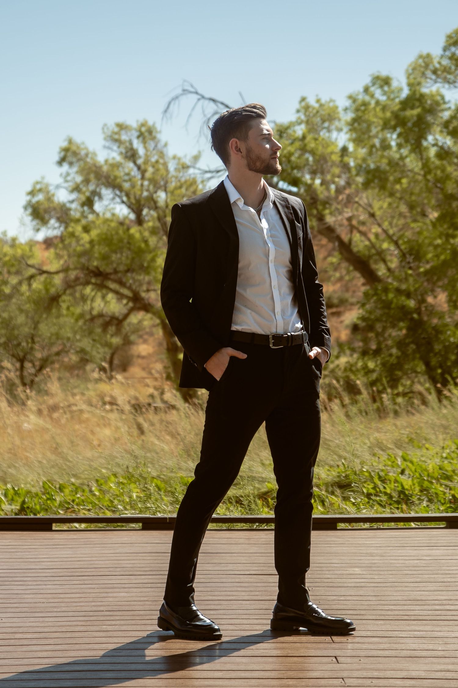 A stylish young man in a black suit and white shirt stands confidently on a wooden deck surrounded by lush greenery.