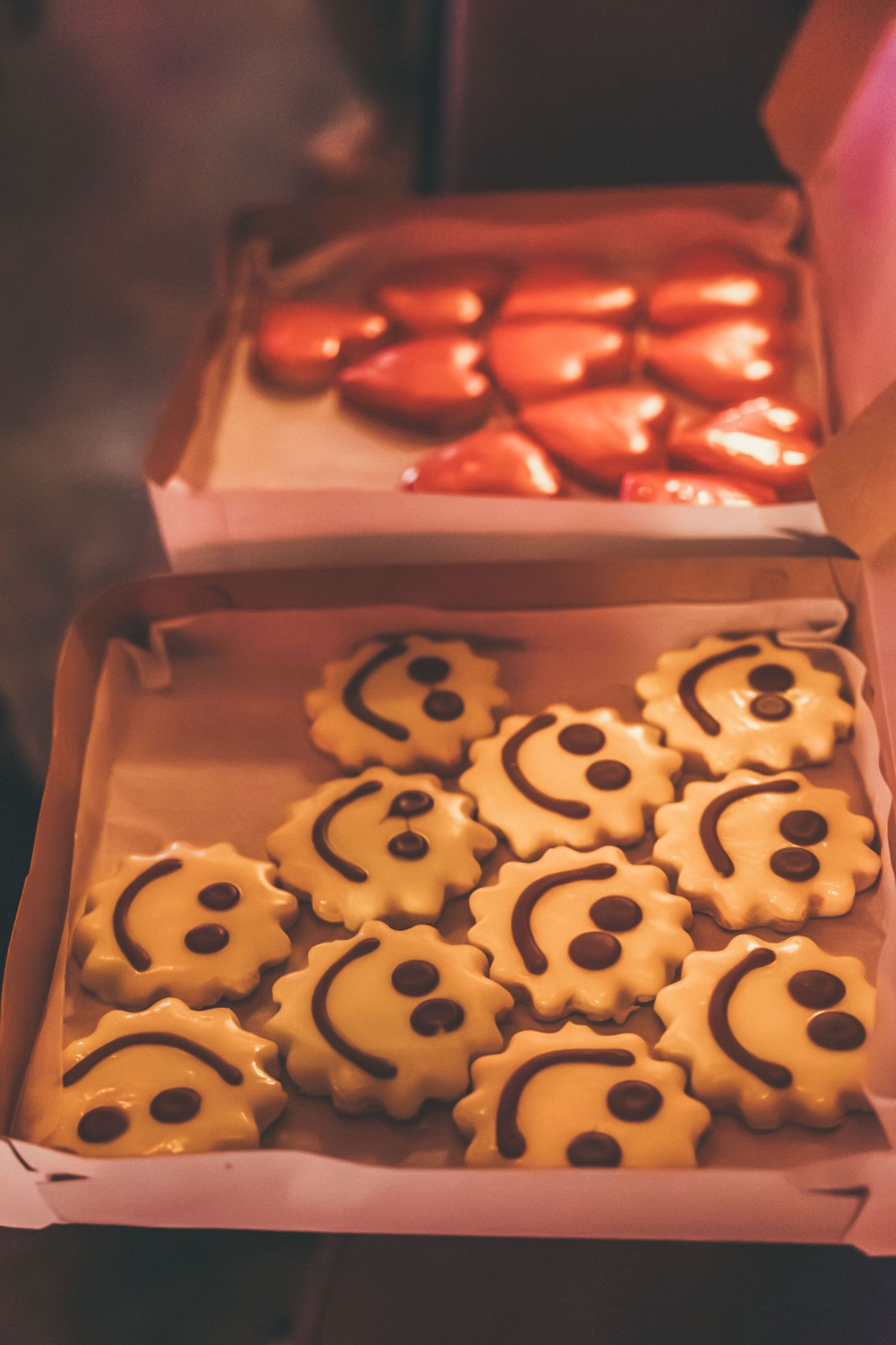 Two open boxes of cookies, one with smiley face decorated sugar cookies and one with heart-shaped chocolates.