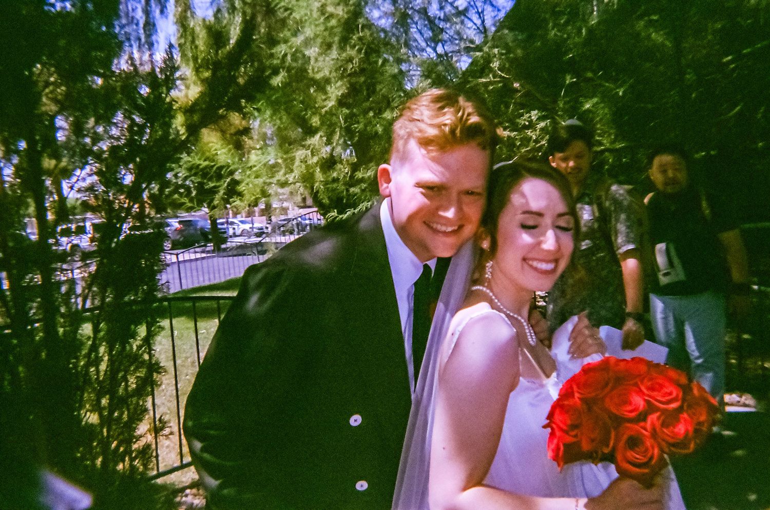A smiling bride holding red roses bouquet stands with groom in black suit outdoors at a sunny wedding ceremony.