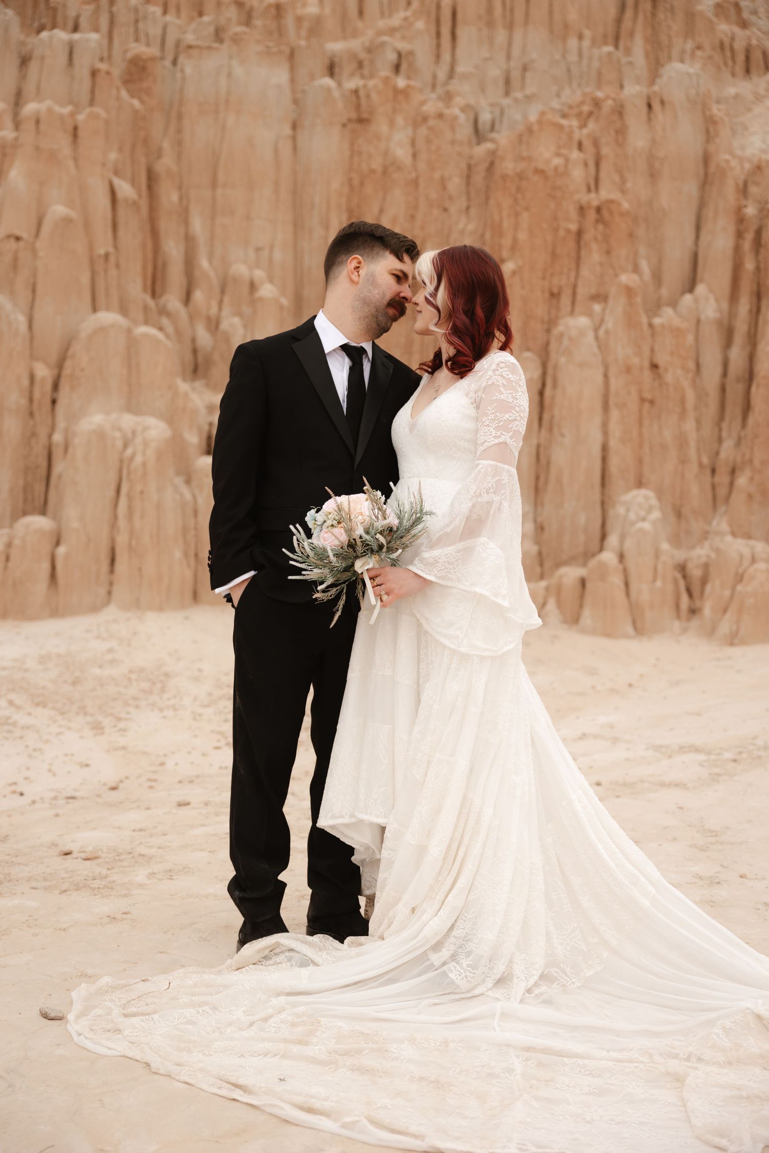 Wedding couple nearly kissing before dramatic sandy rock formations, bride in flowing white lace dress holding bouquet.