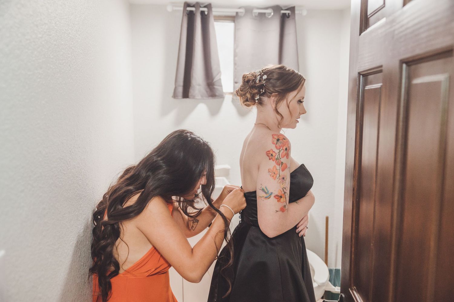 Bridesmaid in orange dress helping bride with black wedding gown in a bridal getting ready room.
