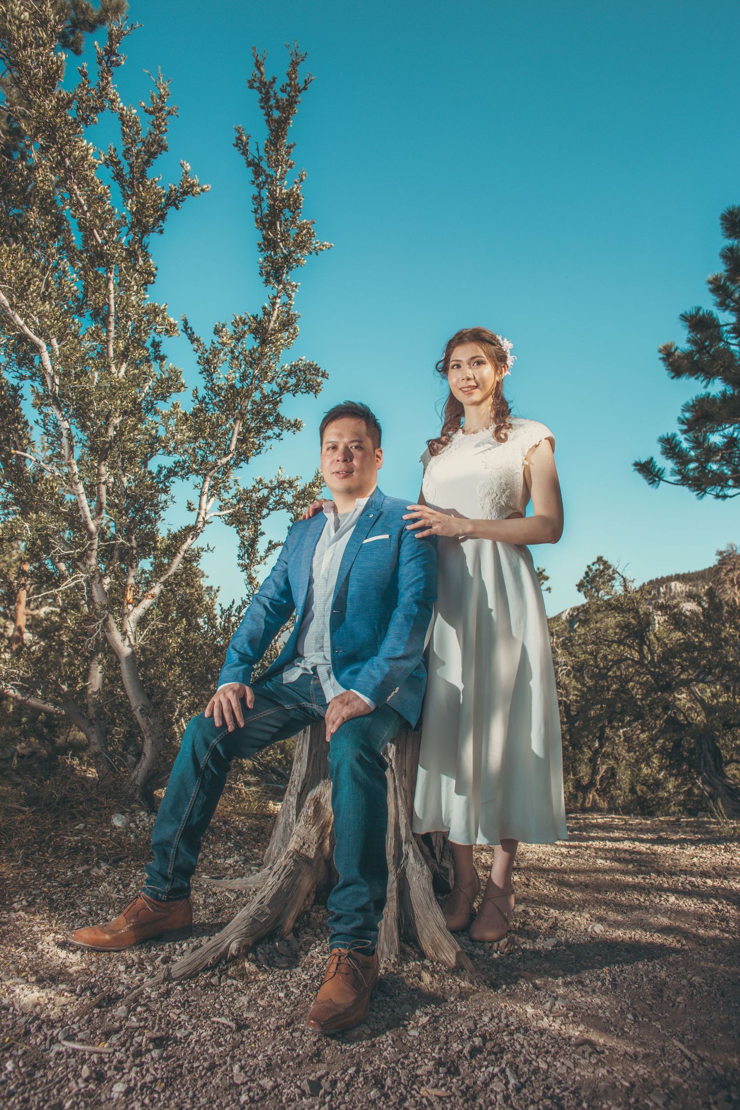 Couple in wedding attire posing outdoors among trees under a clear blue sky, groom seated and bride standing beside him.