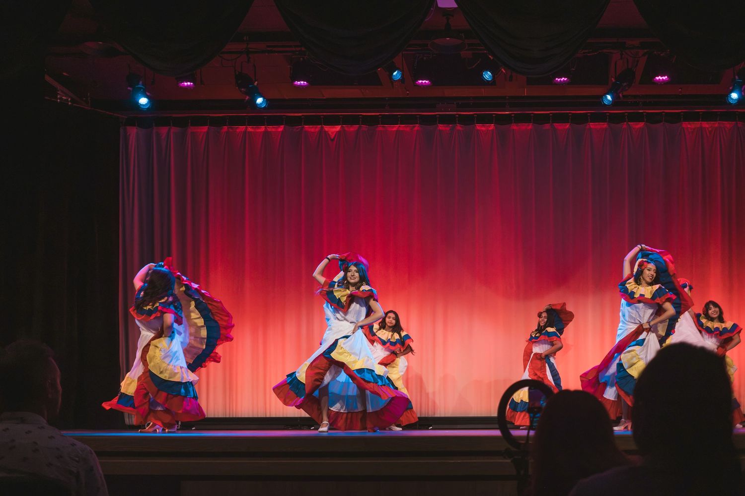 Colorful folkloric dancers perform on a red-curtained stage in vibrant traditional costumes.