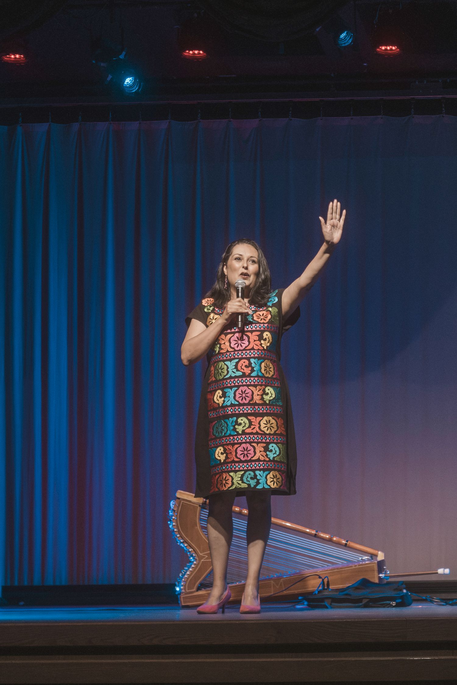 Woman in colorful embroidered dress performing on stage with microphone, hand raised, harp visible in background.