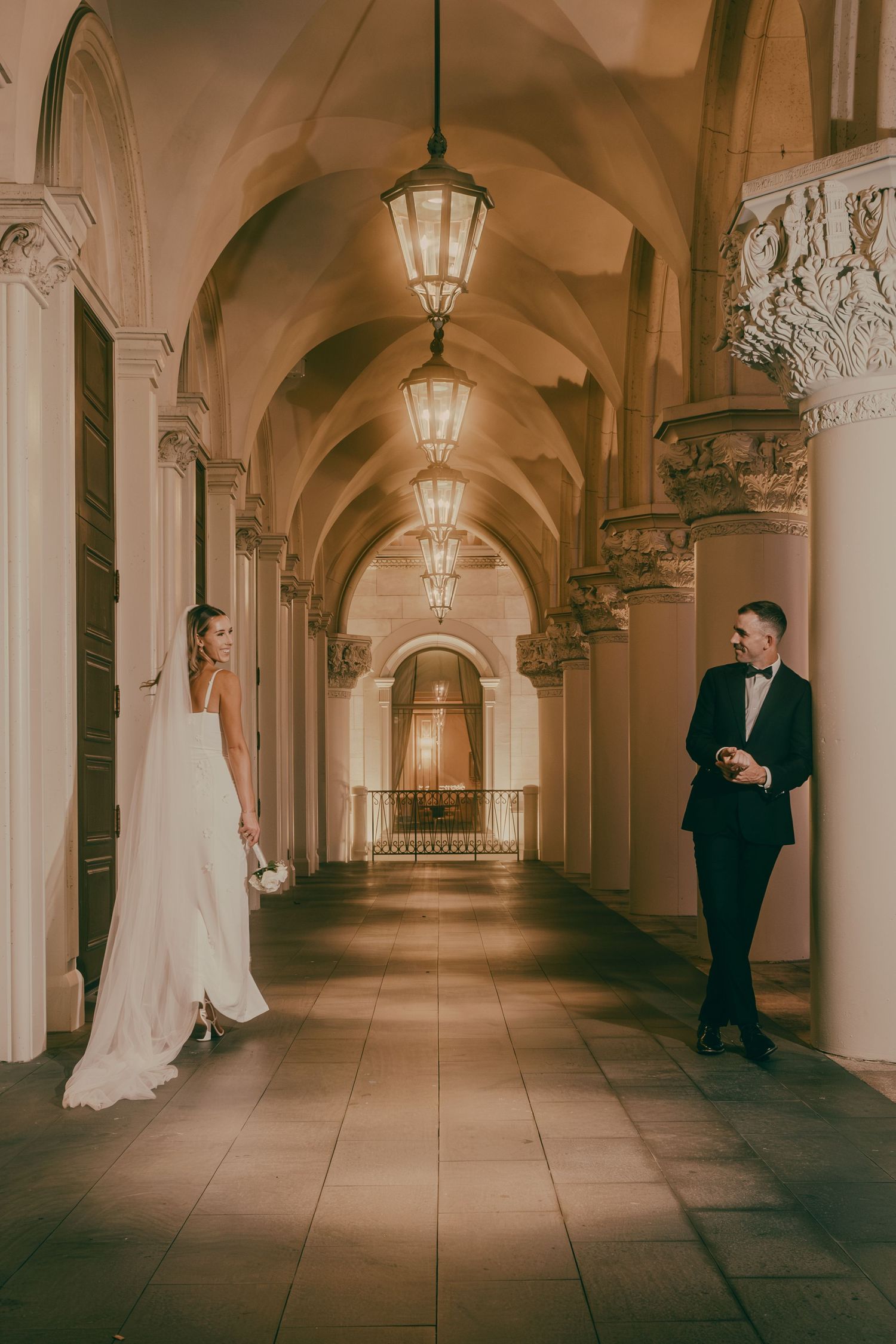 Bride in backless gown and groom in tuxedo pose in warmly lit ornate arched hallway.